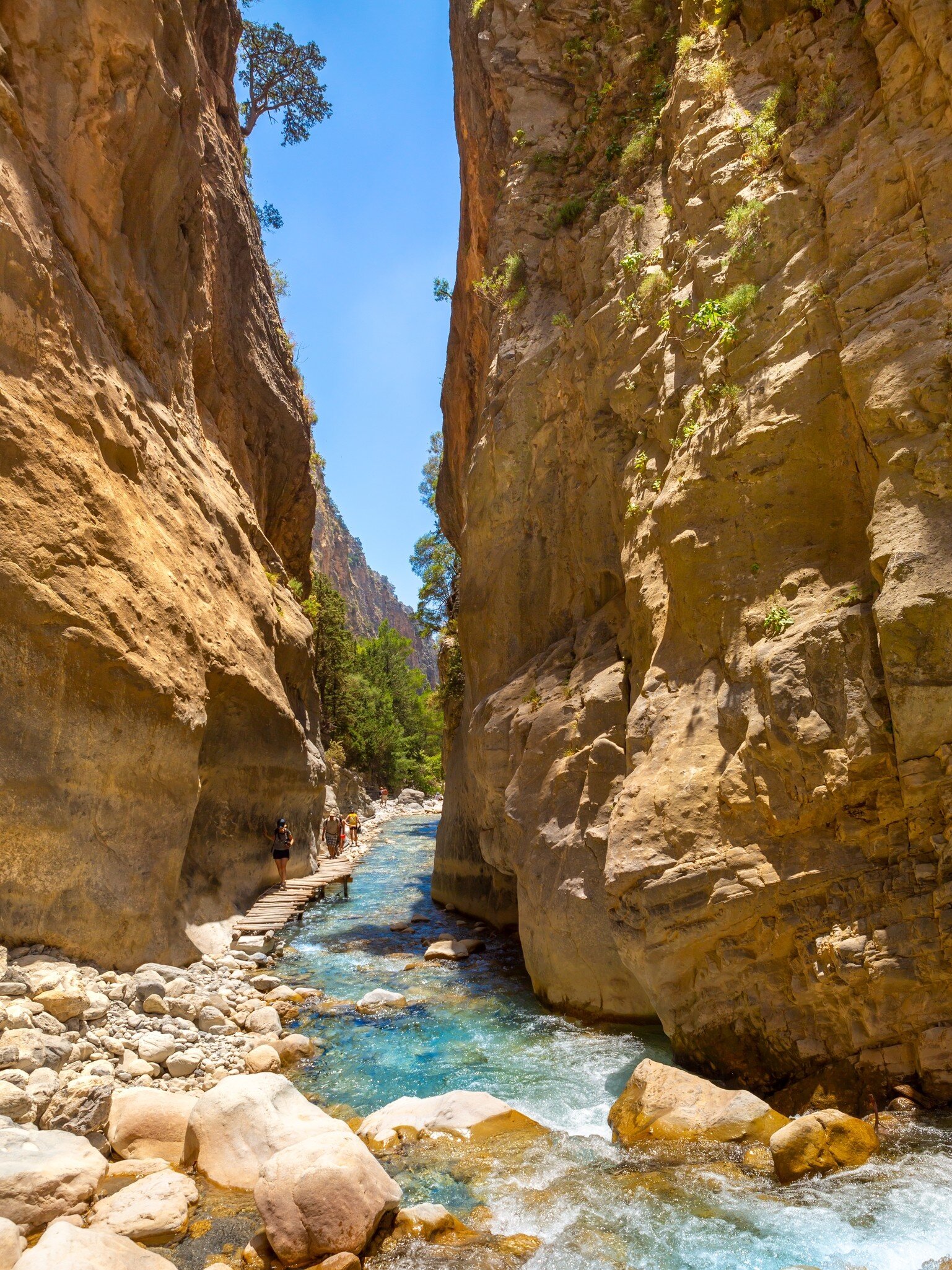 Tourists walk along the clear blue flowing water on the hiking trail in the National Park Samaria Gorge, Crete, Greece.