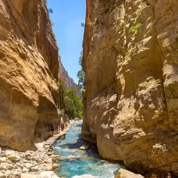 Tourists walk along the clear blue flowing water on the hiking trail in the National Park Samaria Gorge, Crete, Greece.
