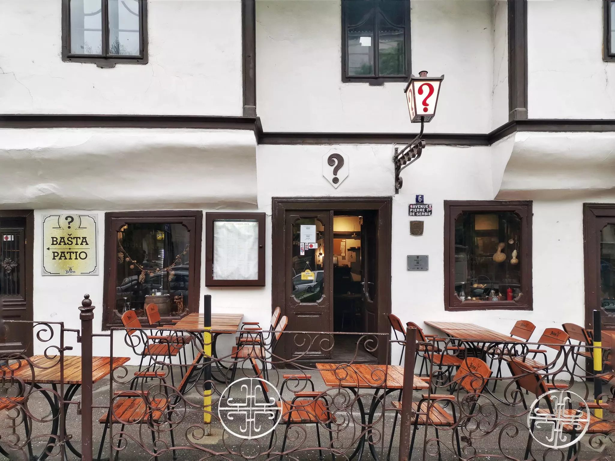 The exterior of a coffeehouse, with tables and chairs on the street. A large lamp with a red question mark marks the entrance.