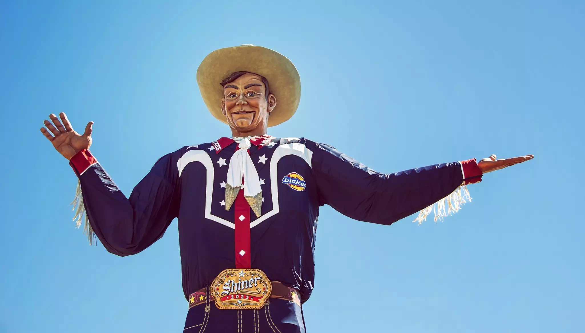 Large statue of a cowboy dressed in blue clothing with white stars, red collar and cuffs, white fringe along the arms, and a cowboy hat on a sunny, cloudless day.