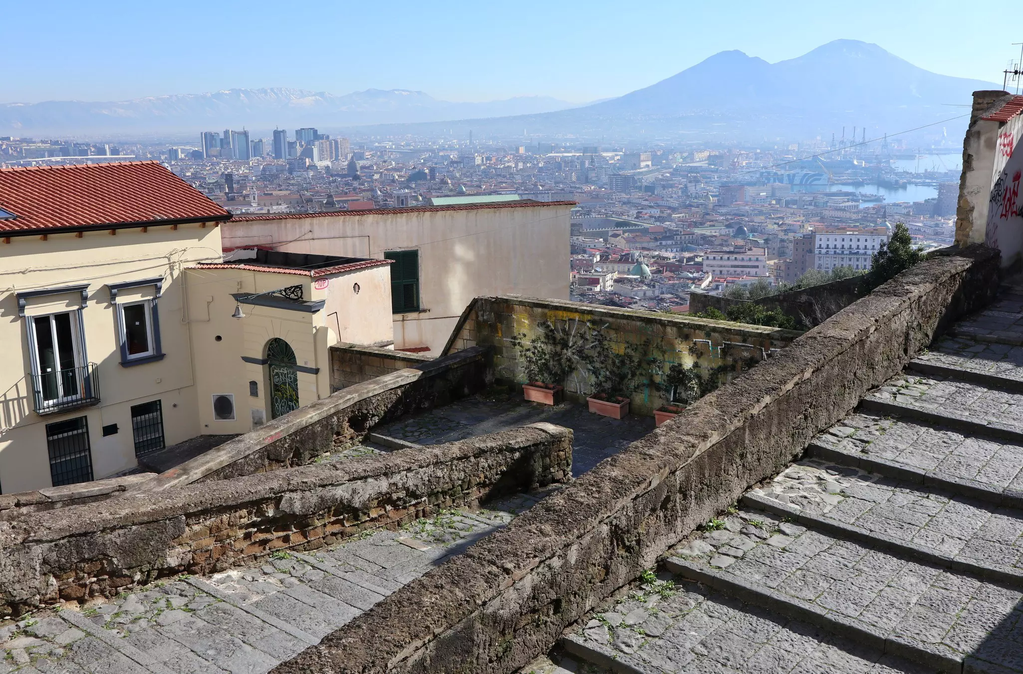 A staircase made of stone leads down a hill toward the center of a city. A volcano is visible in the distance.