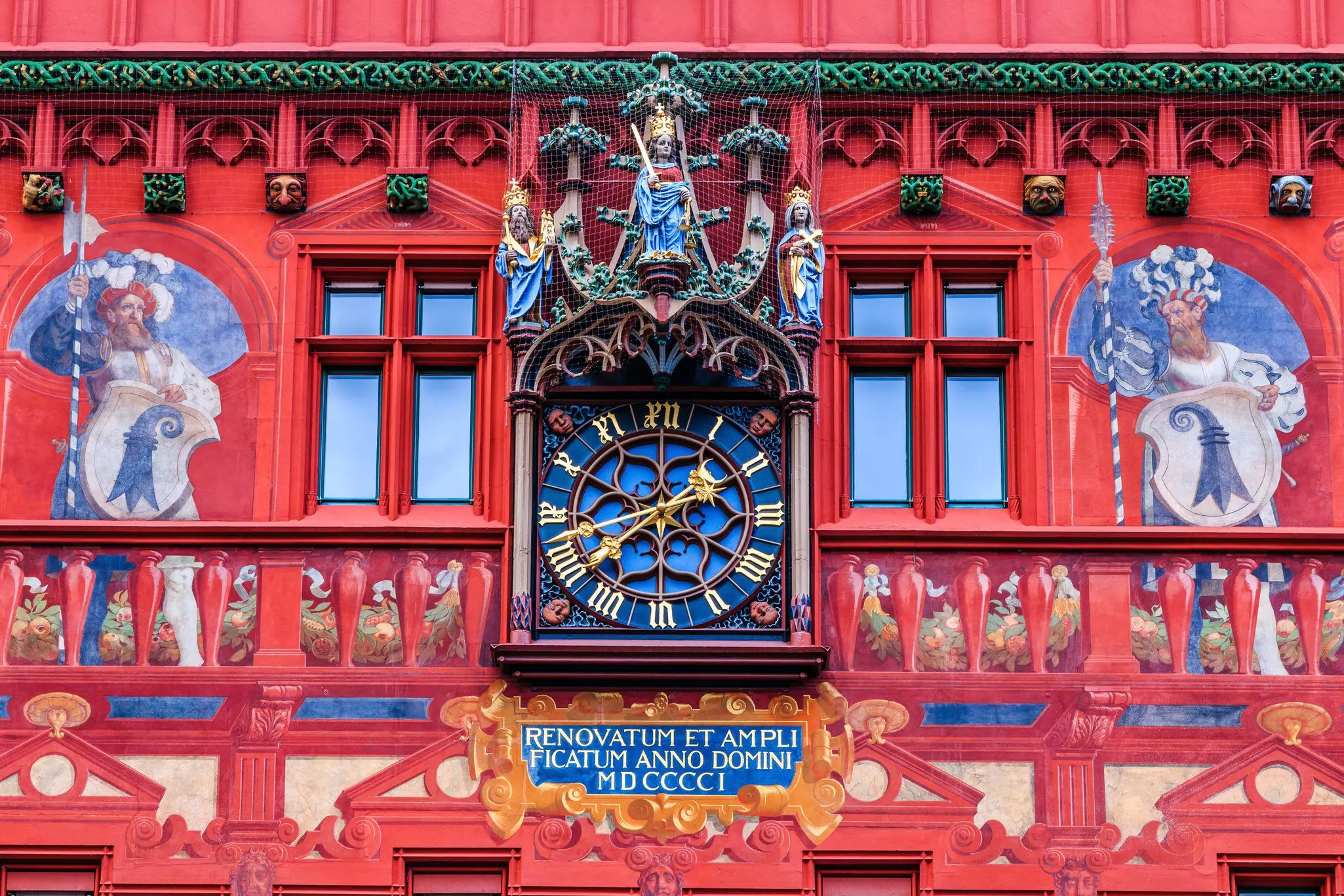 An elaborate clock with a blue face and golden numbers and hands on a richly decorated red building in Switzerland.
