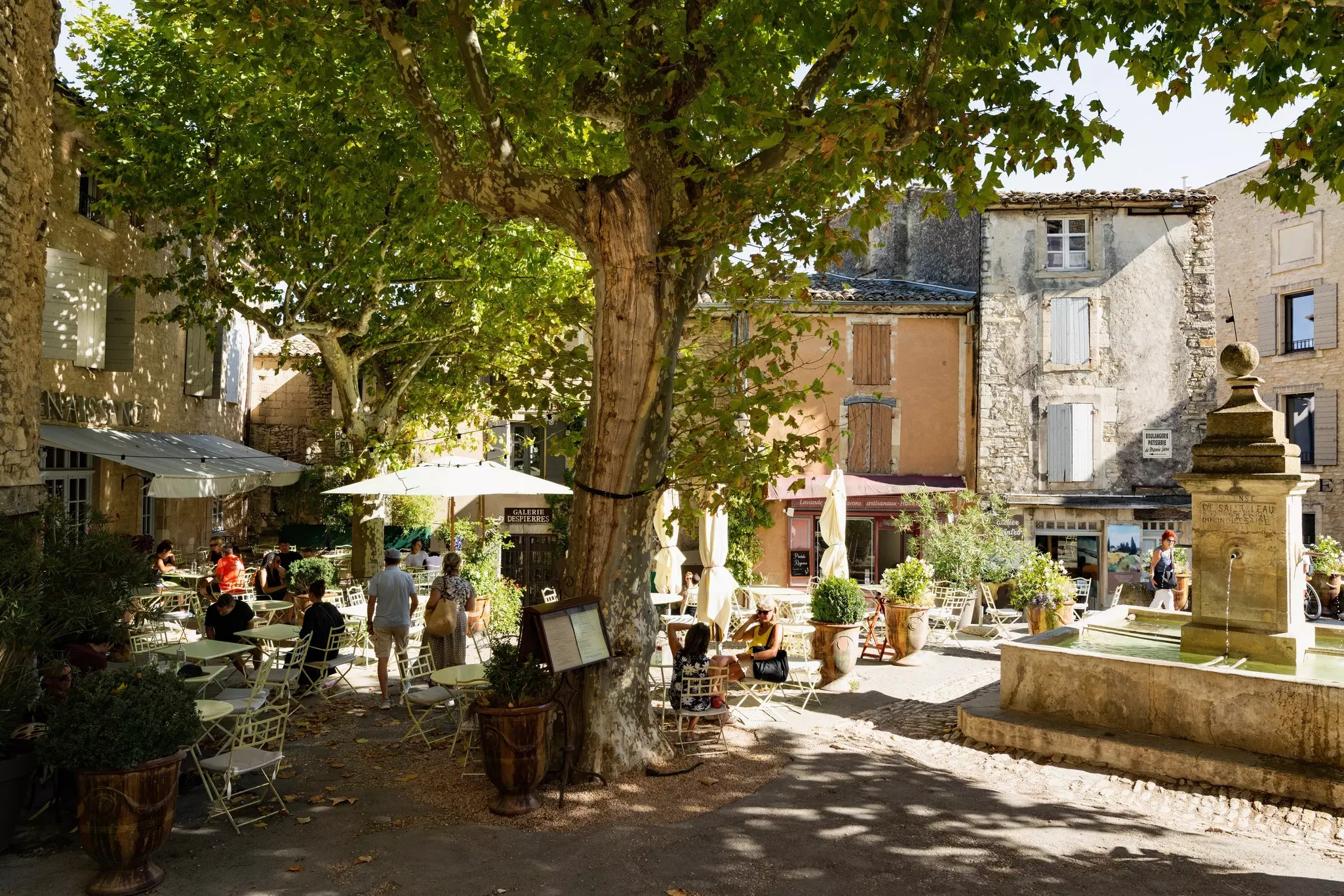 A medieval square in the heart of the village of Gordes, France, full of large trees and with a fountain at the center.