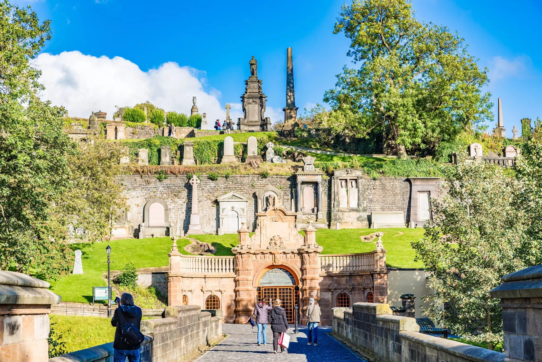 People walk towards a large hillside cemetery with distinctive monuments in Glasgow, Scotland