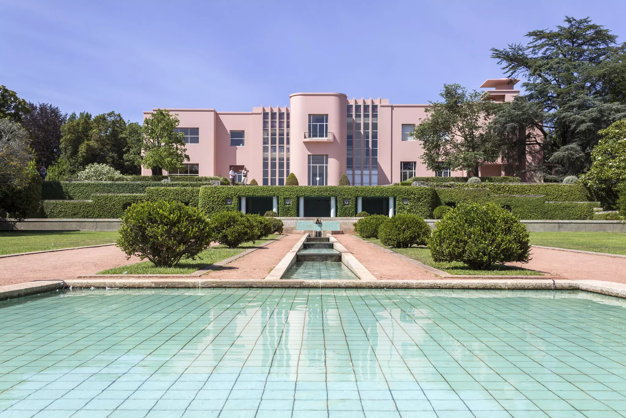A pink house in the art deco style surrounded by landscaped fountains and gardens 