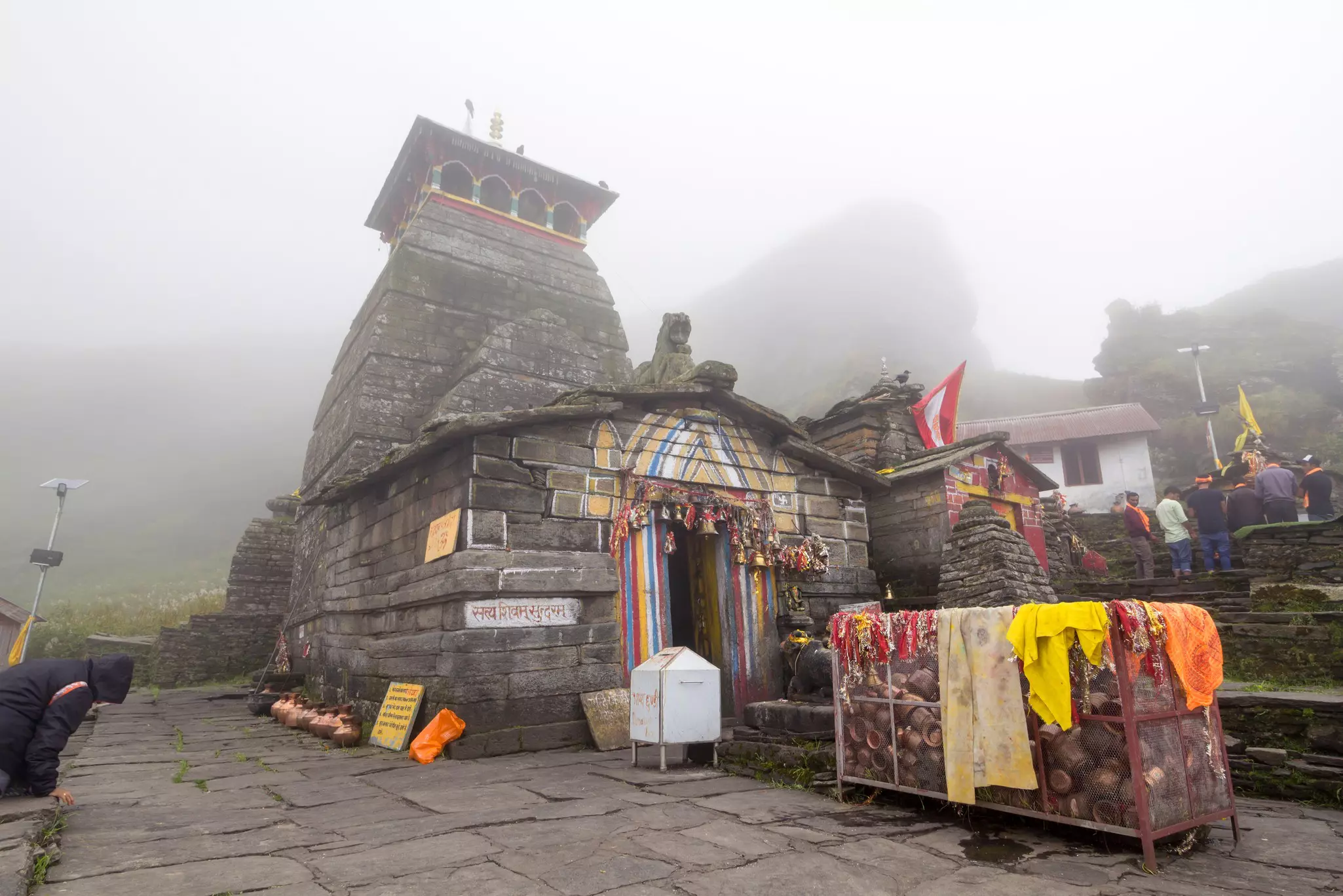 A stone temple with colorful painting on the doorway surrounded by flagstone patio. A man kneels in the foregournd and other people gather in the distance on a misty day.