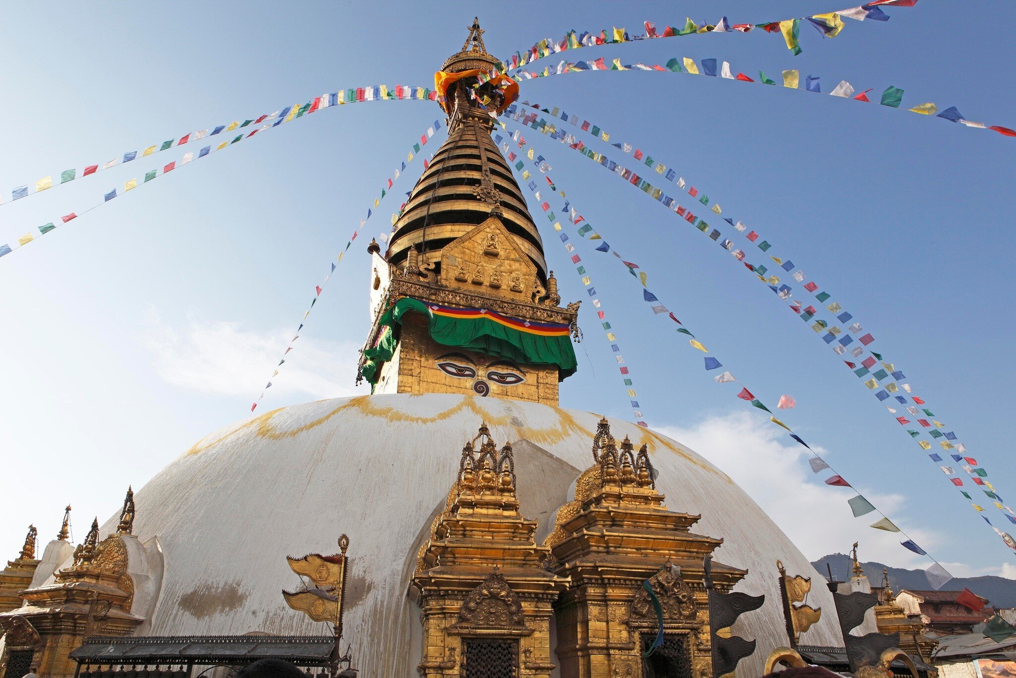 White domed stupa with painted eyes on a gold tower, strings of colourful prayer flags against a blue sky.