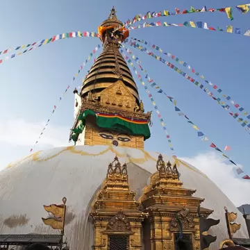 White domed stupa with painted eyes on a gold tower, strings of colourful prayer flags against a blue sky.