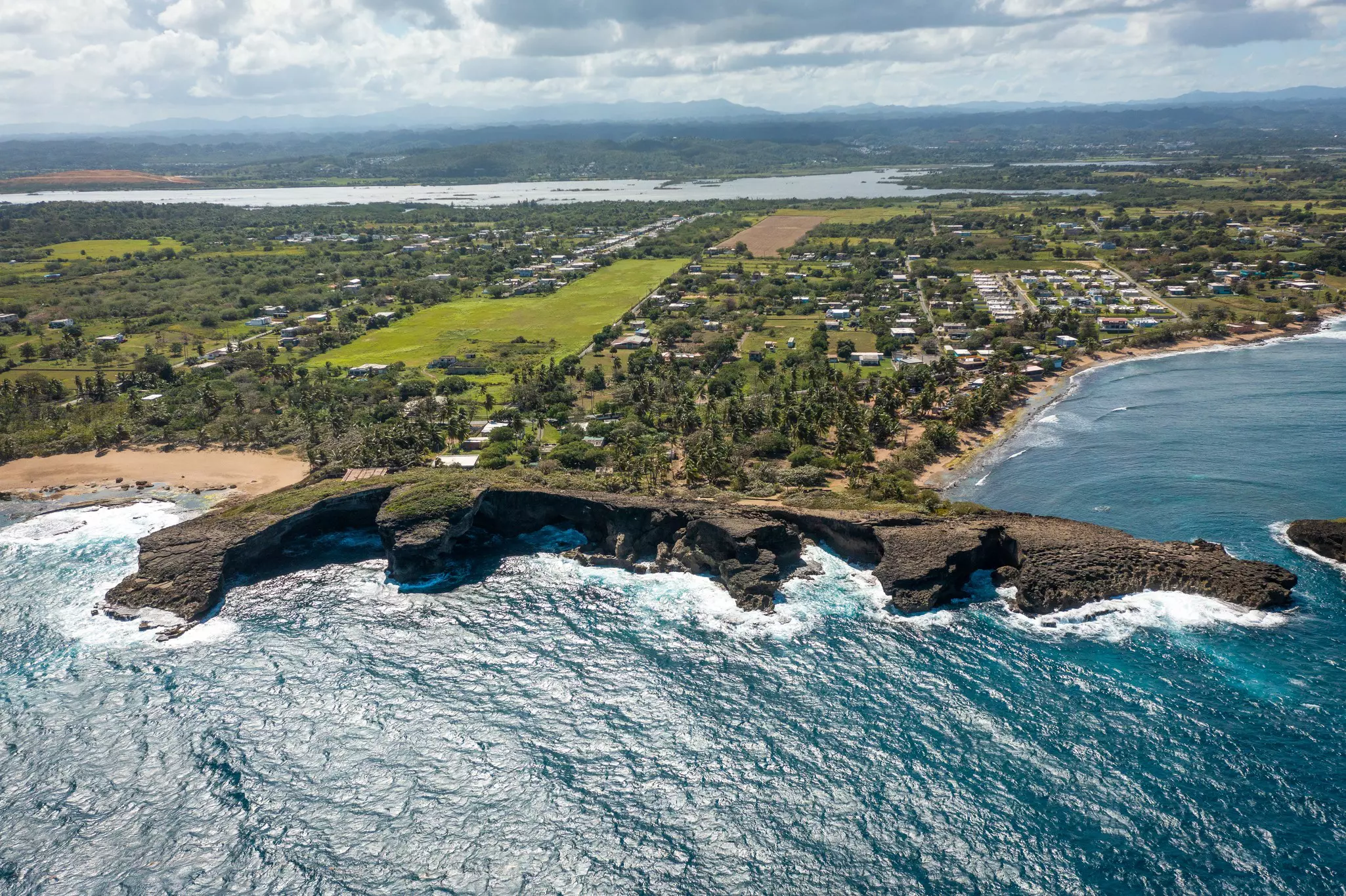 Aerial view of rocky outcropping with grass, trees and a town in the distance and the ocean in the foreground on a bright, sunny day.