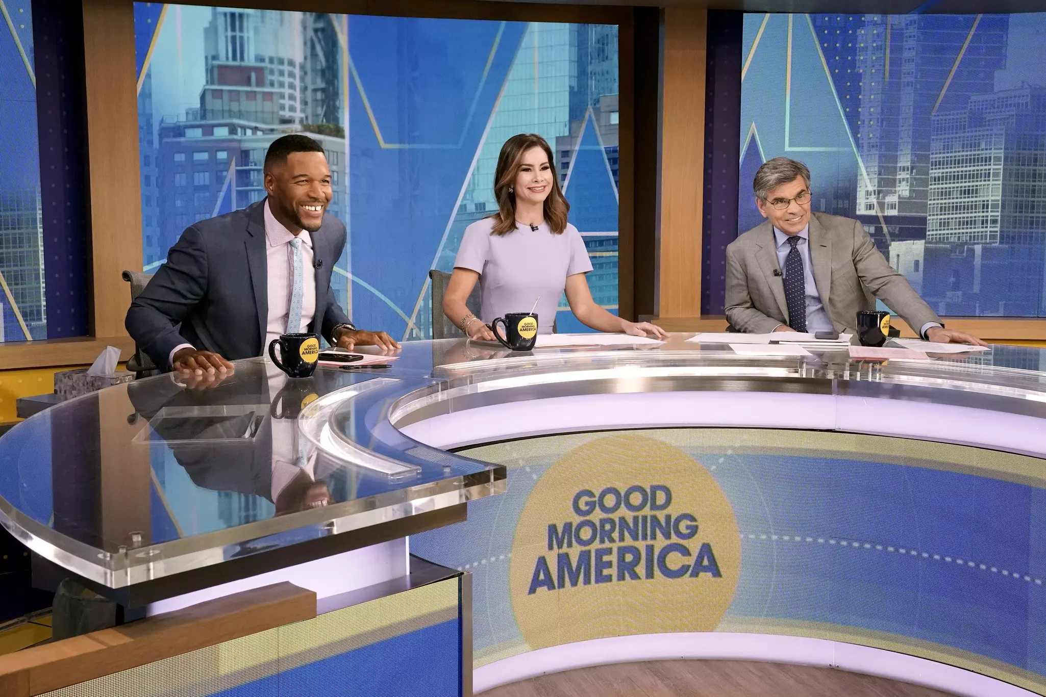 Three hosts, Michael Strahan, Rebecca Jarvic, and George Stephanaopoulous, sit at a curved desk with the words "Good Morning America" on it and smile toward a camera that is offscreen.