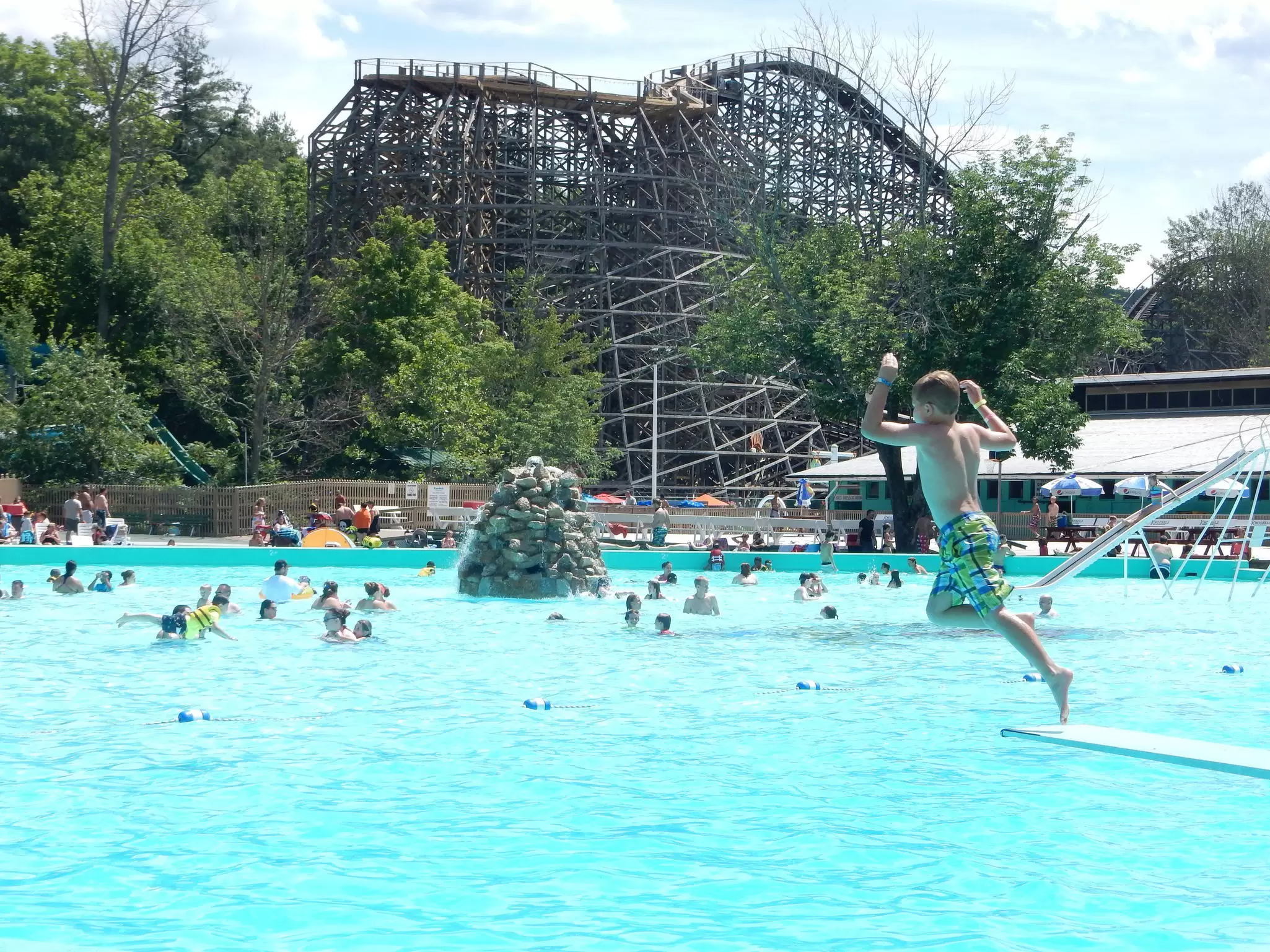 A kid jumps into a large swimming pool with a wooden roller coaster nearby at Knoebels Amusement Park, Elysburg, Pennsylvania