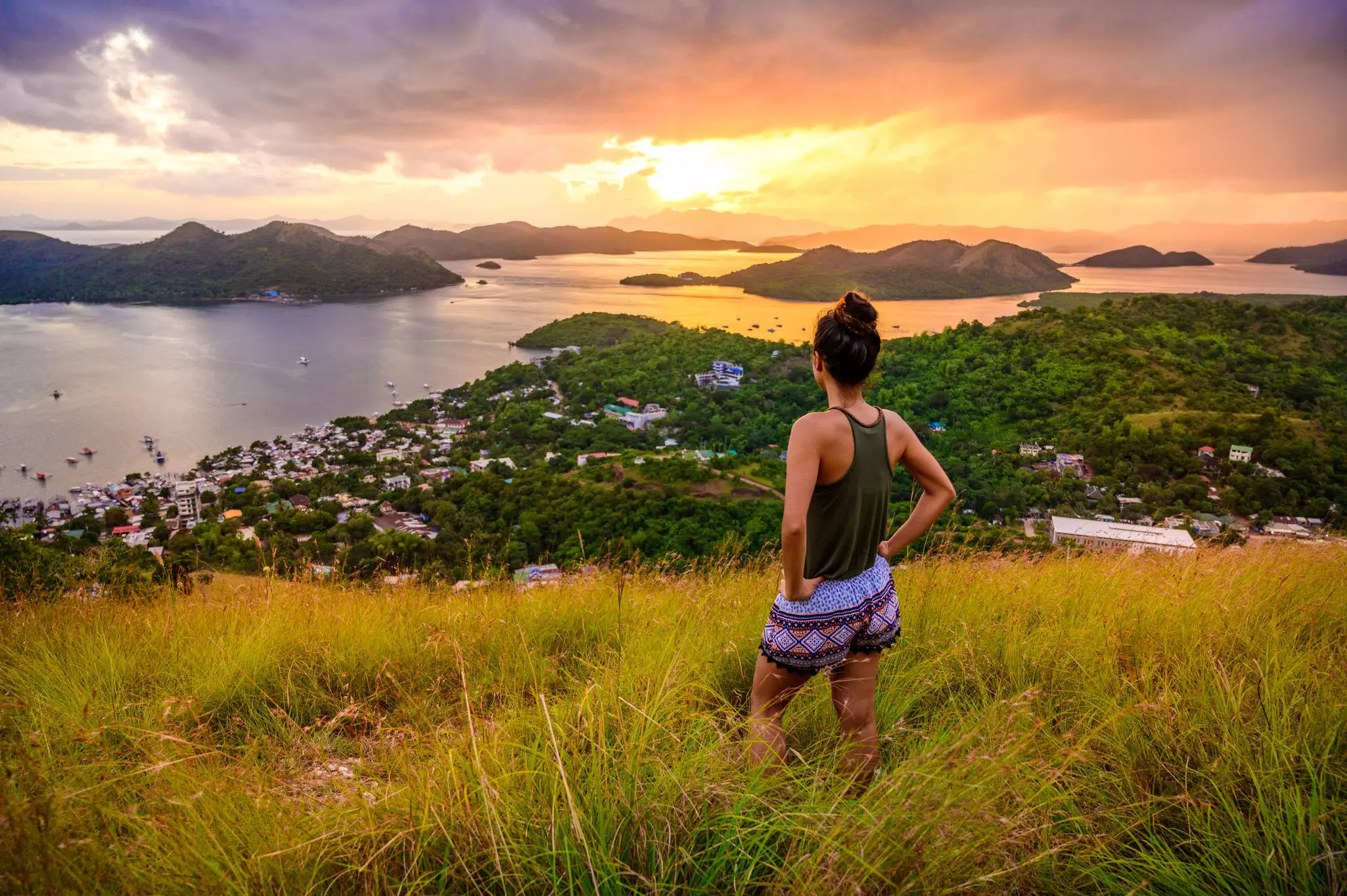A woman is seen from behind standing in a grassy area on a hillside looking out the sunset, at a village and hilly islands beyond