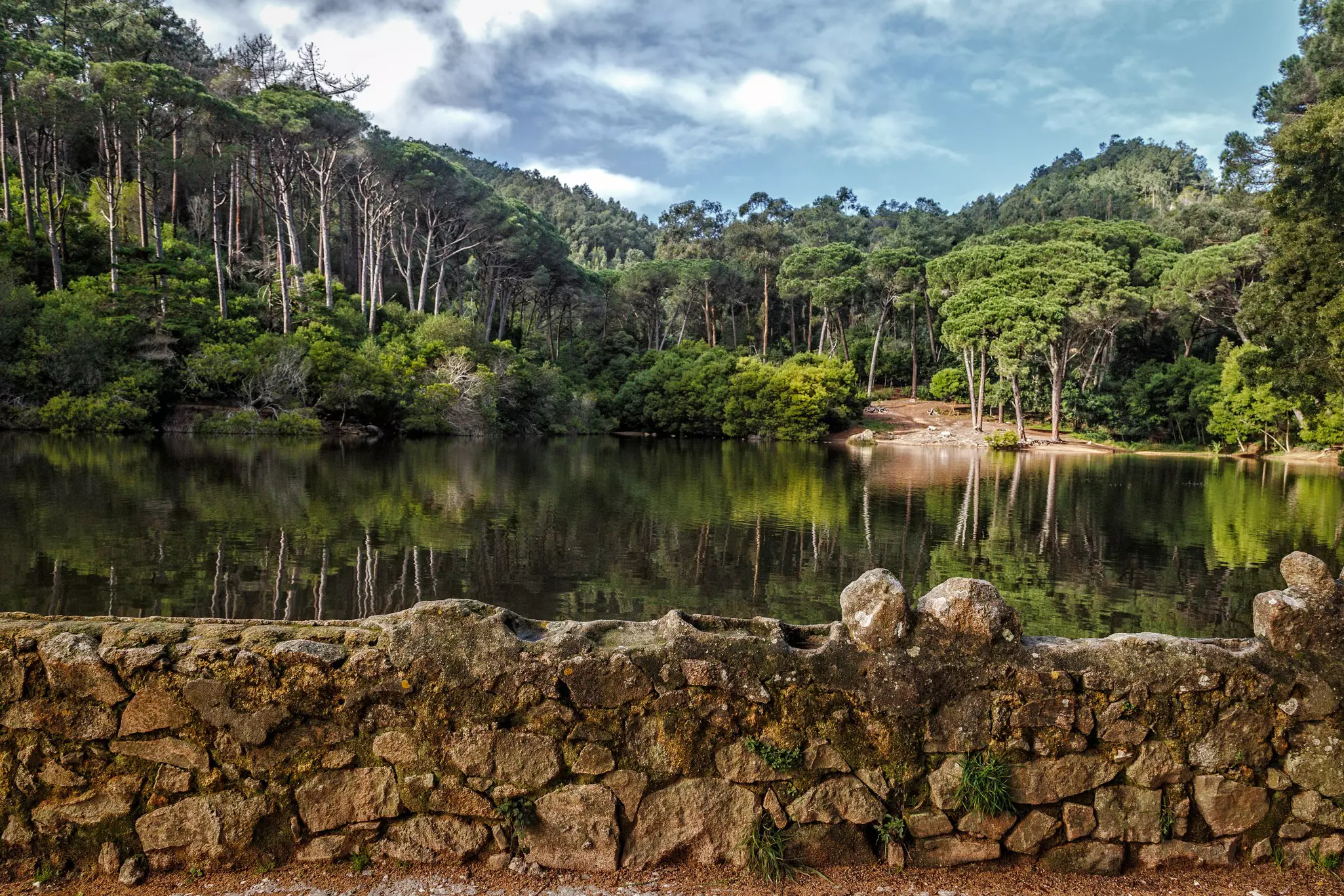 A rock wall is seen in front of a smooth lake, with abundant trees surrounding it and rising along the nearby hillsides
