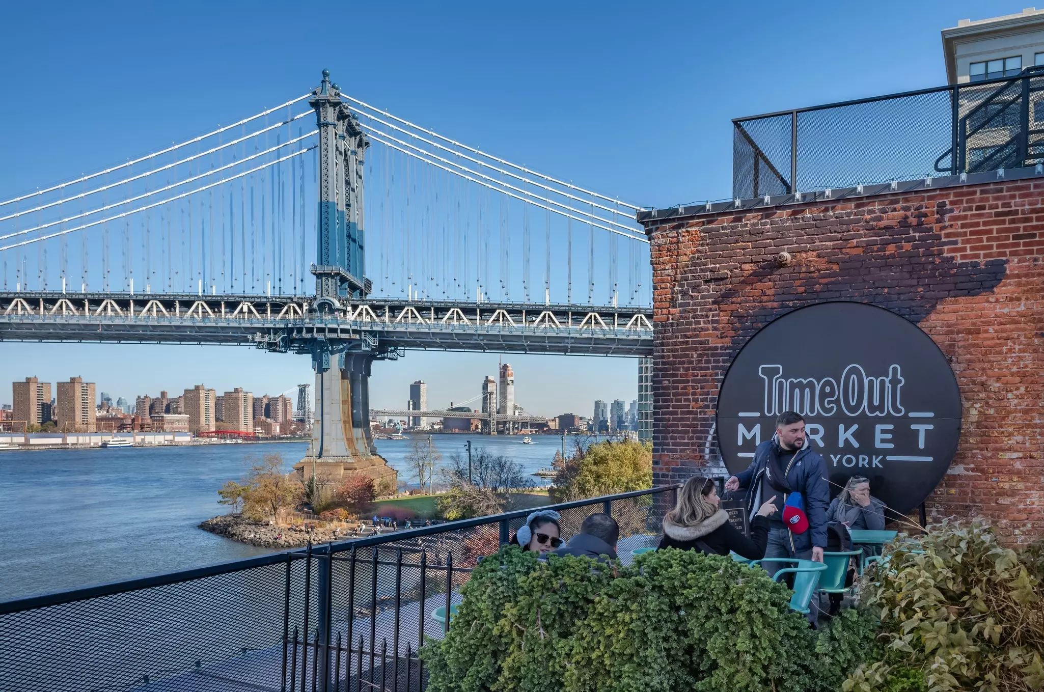 Table and chairs on an outside terrace, filled with diners. In the background is the river and views of Manhattan.