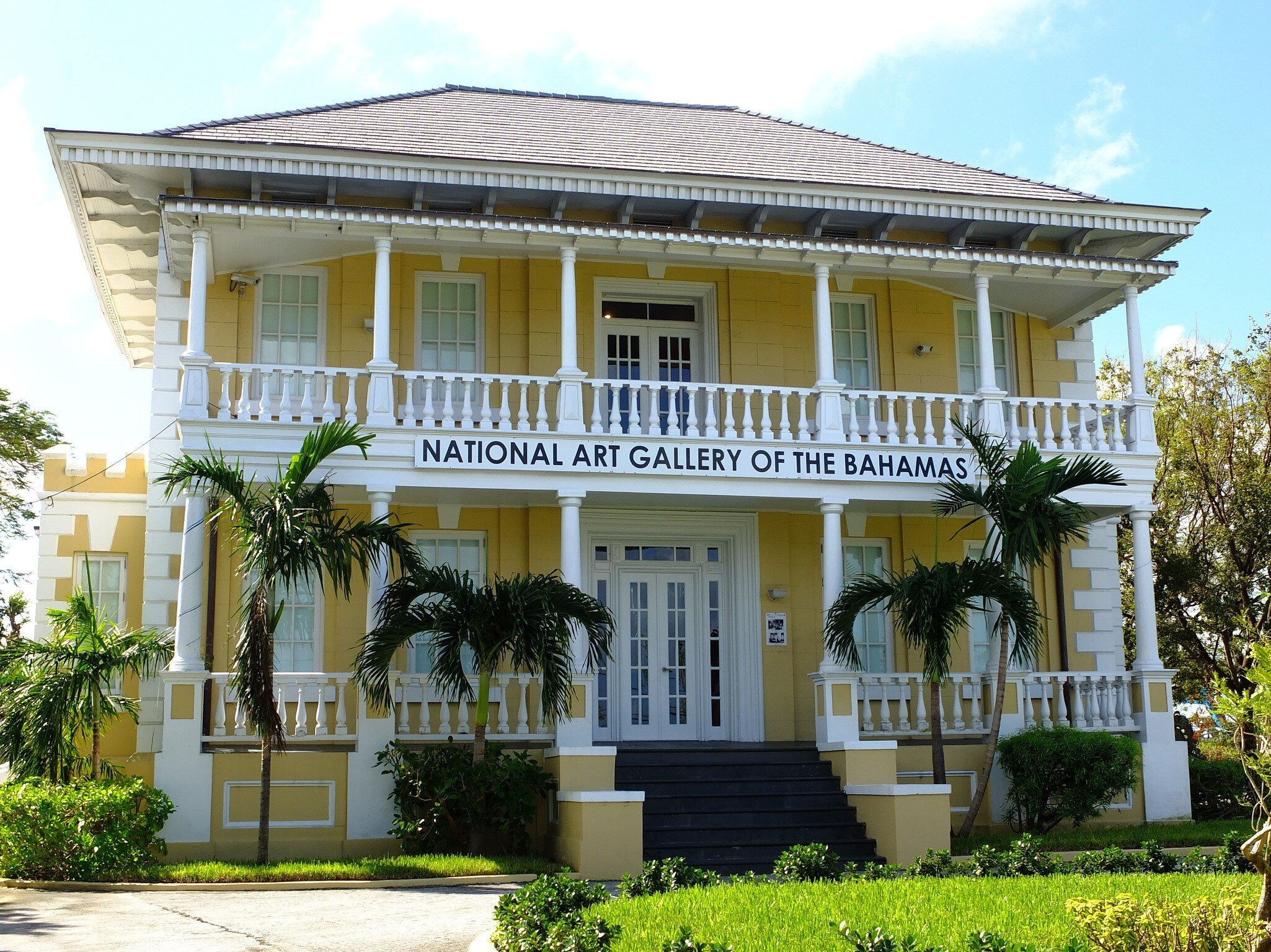 A yellow colonial mansion with white columned verandas on two levels, fronted by palms and a lawn