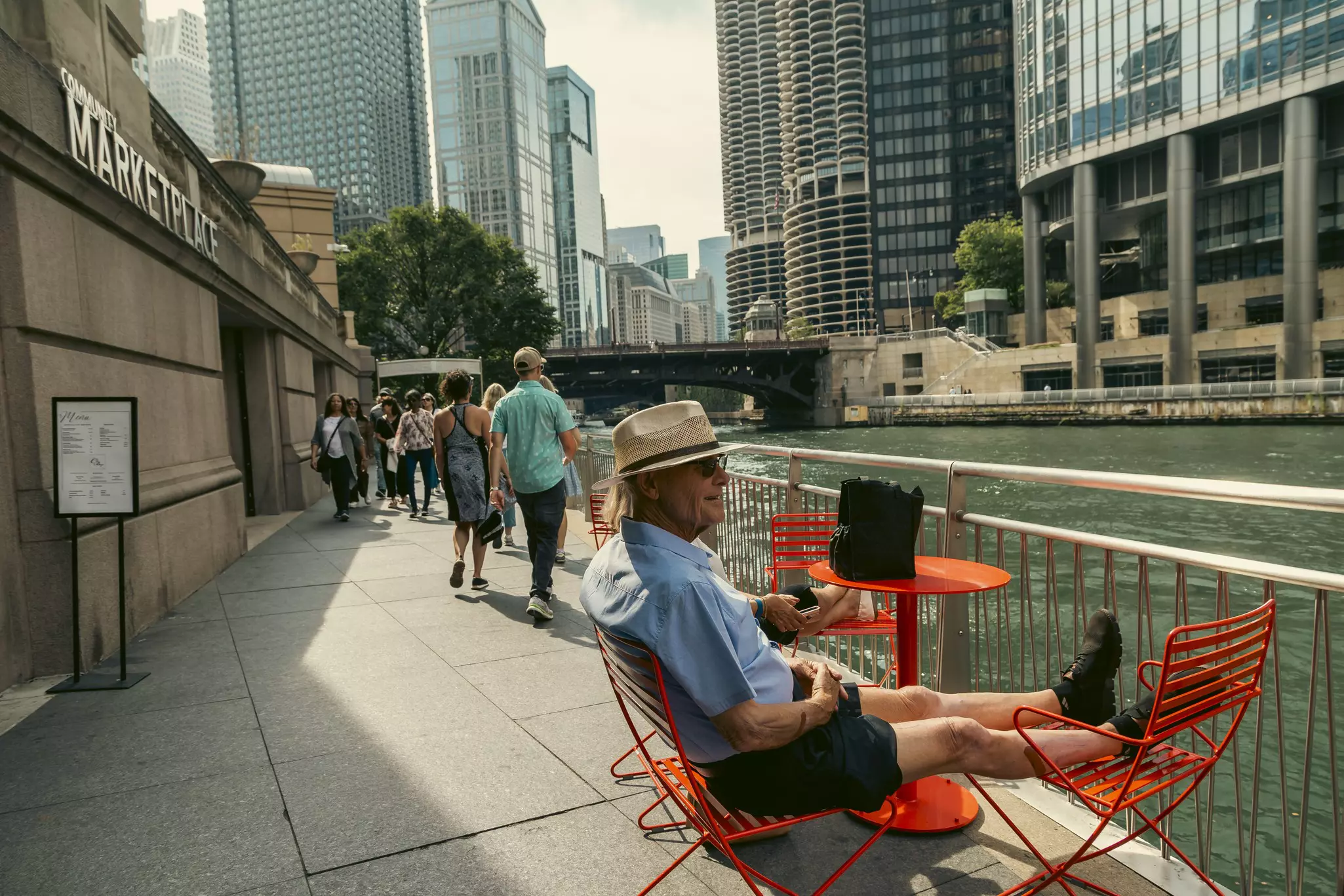 A man sits and puts his feet up on red chairs placed on a walkway along a river in a major city.