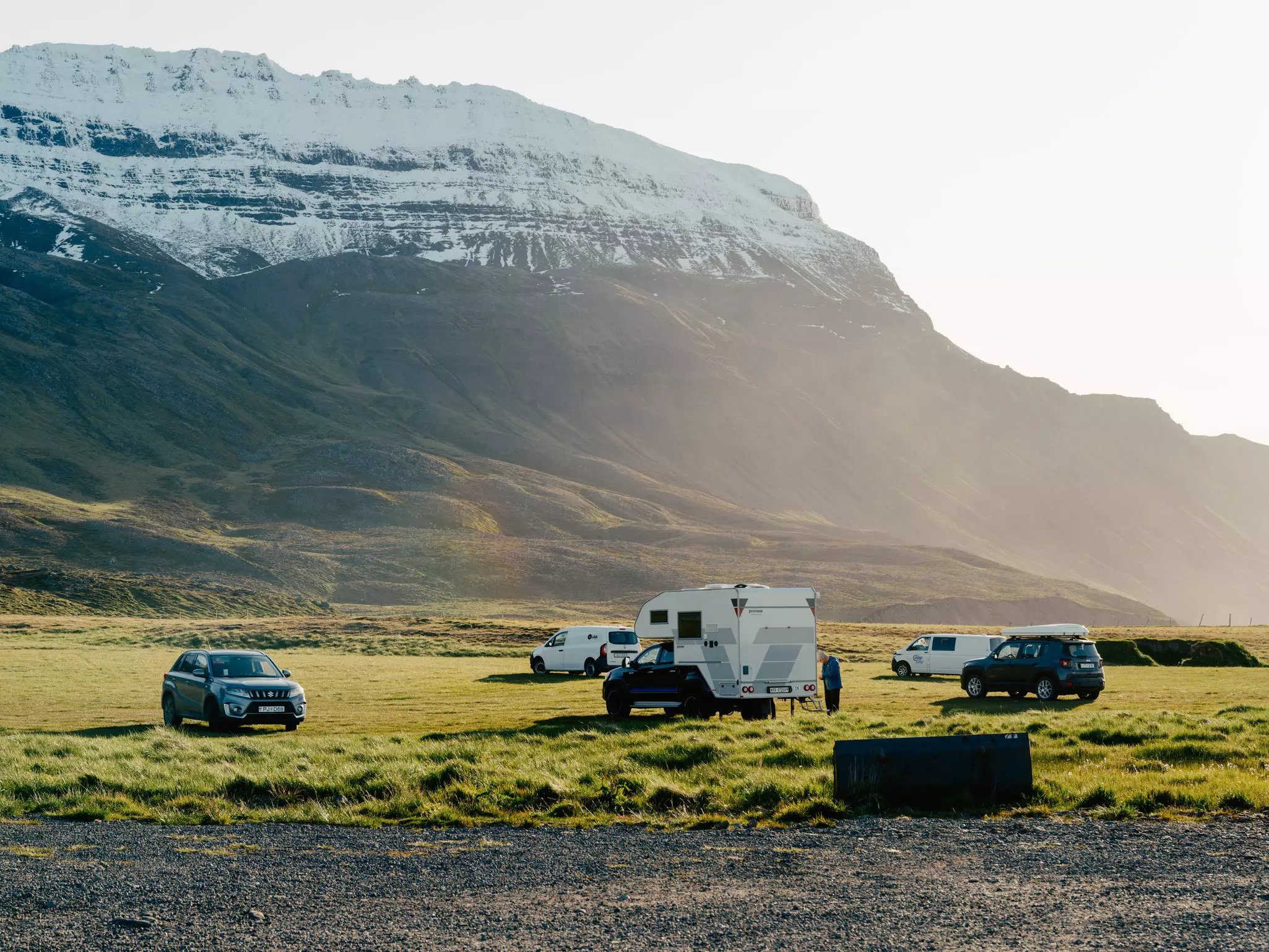 An RV, camper vans and SUVs in a grassy field with snow-capped mountain in the distance near the end of a sunny day.