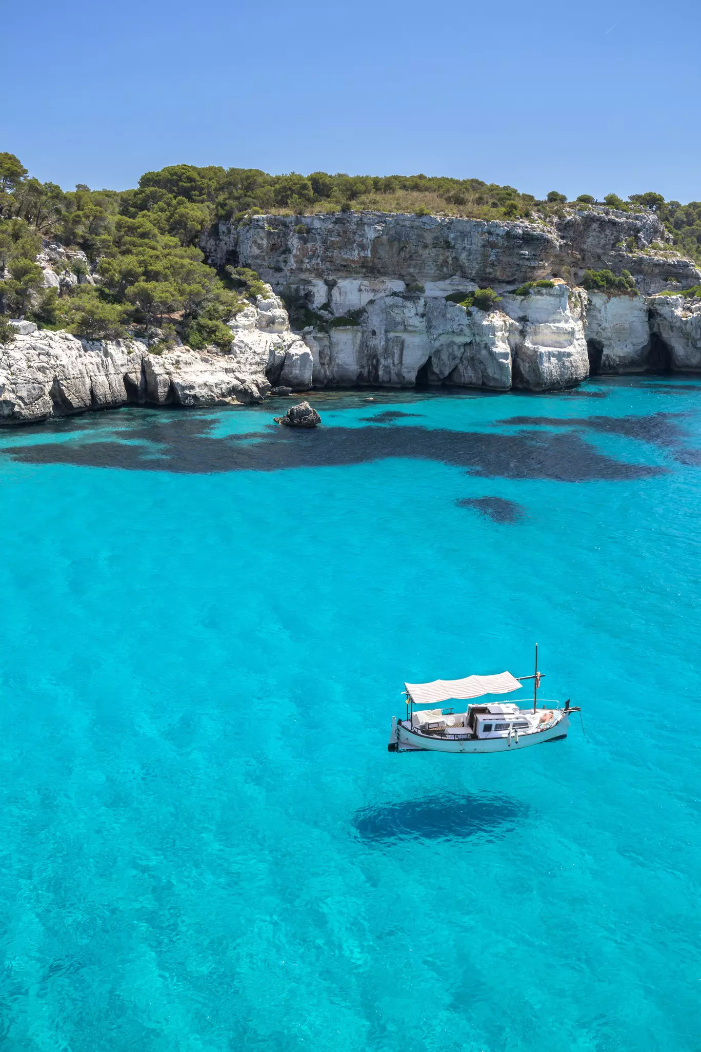 A small sail boat sits in pristine clear turquoise waters near a rocky coastline