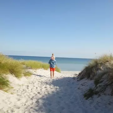 An adult with a child on their shoulders walks on a sandy dune toward blue sea.