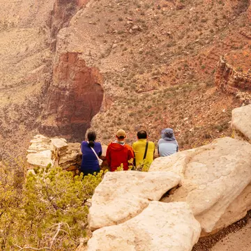 Hikers in colored coats sit on the rocky canyon rim in Grand Canyon National Park, USA.
