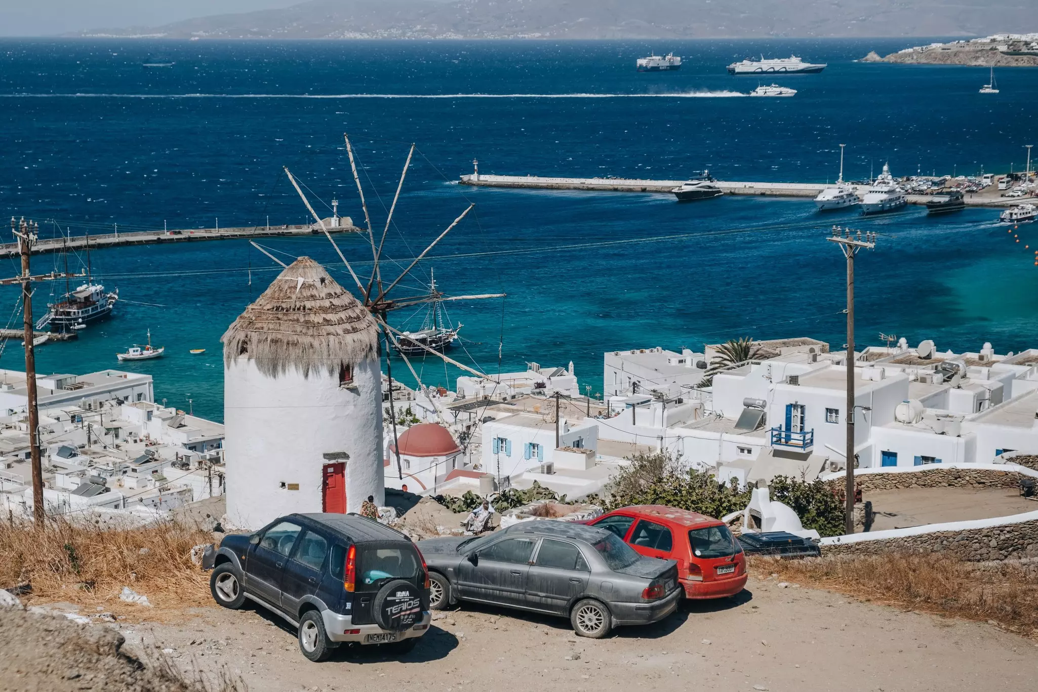 Three cars parked on a dirt track near a windmill on a hill above a small harbor town.