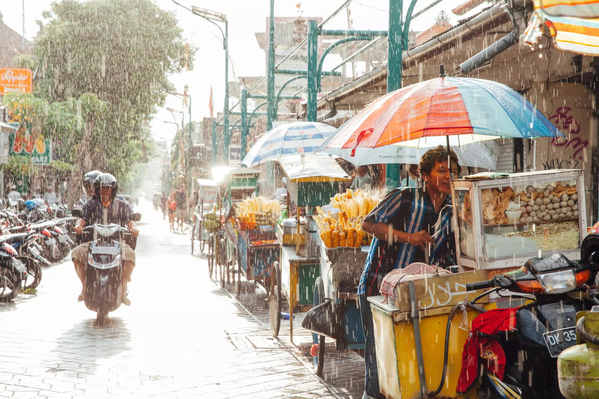 A motorcycle with two passengers drives down a narrow street as the rain pours down. A nearby street-food vendor takes shelter under an umbrella.