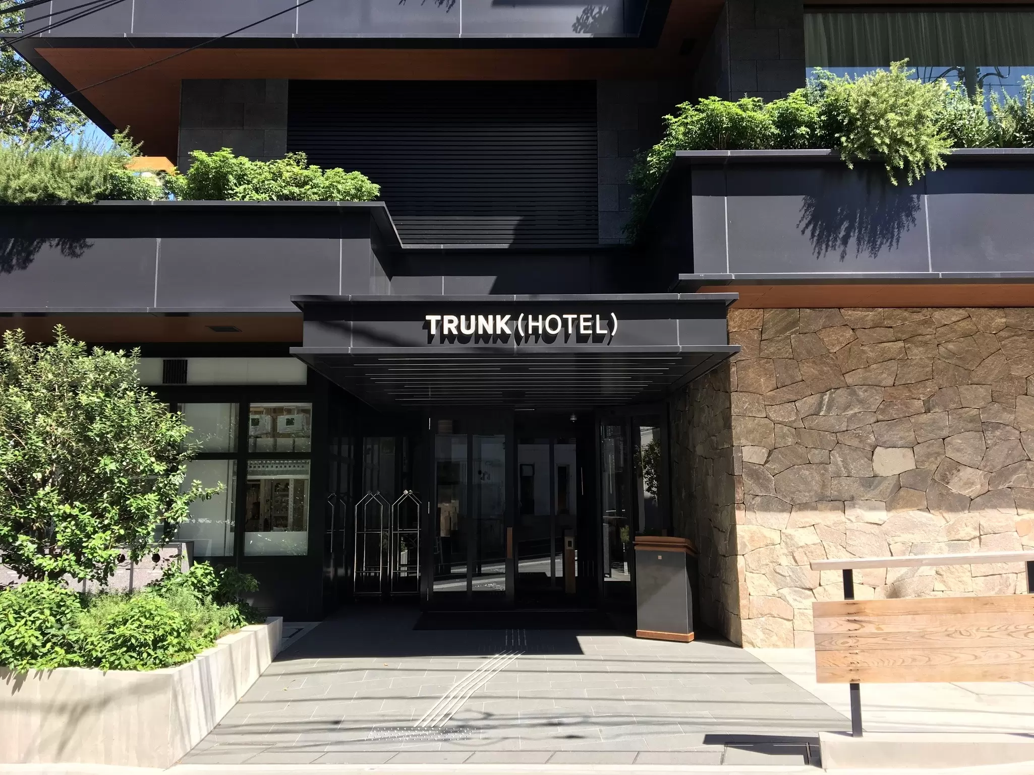 Entrance of a black building with green plants nearby. The words "Trunk Hotel" are over the glass doors.