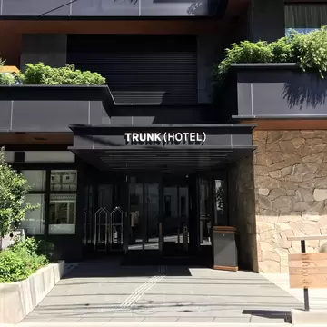 Entrance of a black building with green plants nearby. The words "Trunk Hotel" are over the glass doors.
