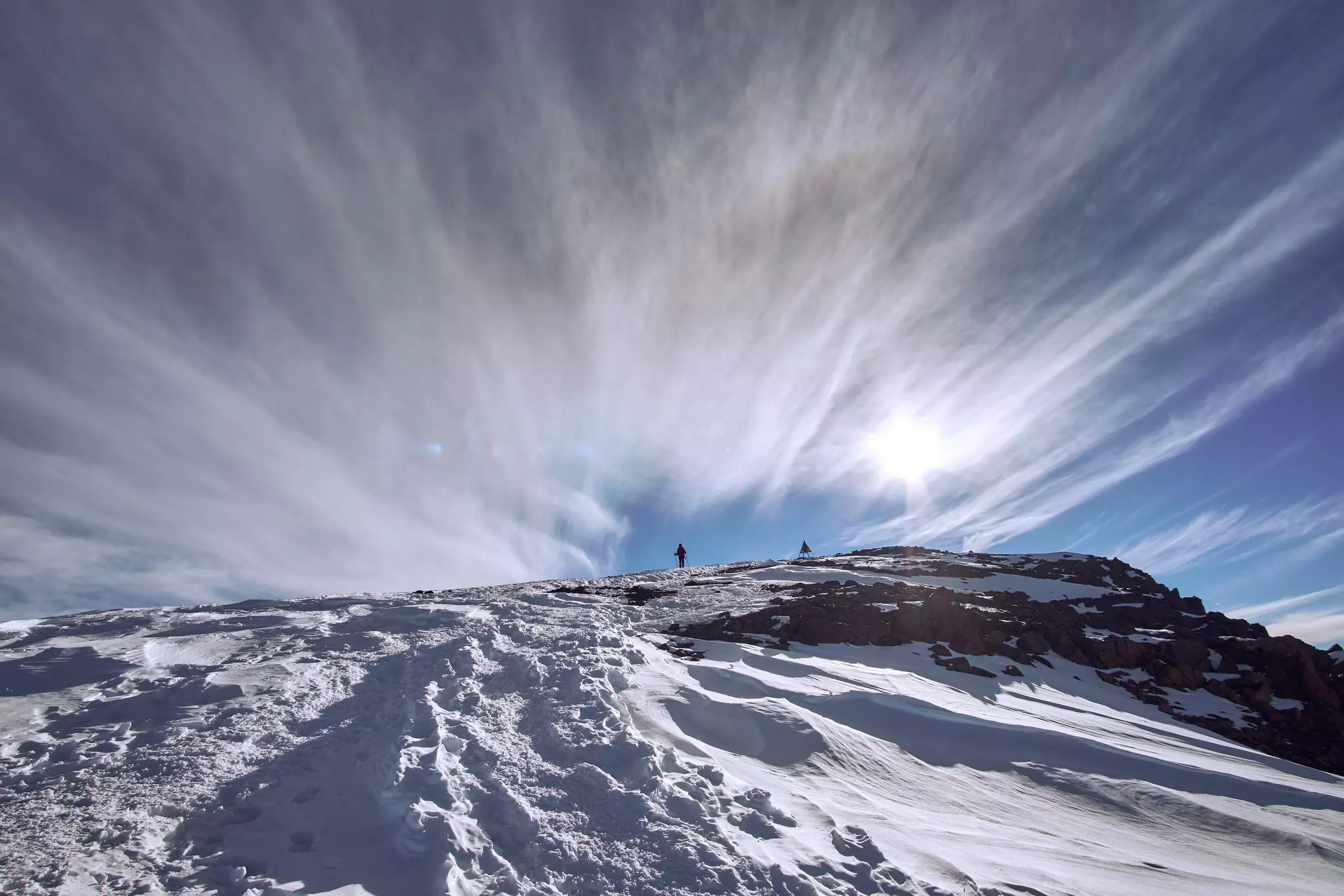 This image looks up a path in the snow towards a triangular cairn at the summit of Jebel Toubkal; a climber is seen approaching the top. The blue sky has wispy clouds.
