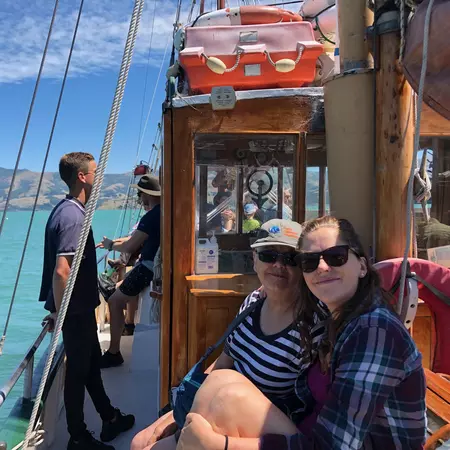 Two women sit on the deck of a wooden sailing ship with a turquoise sea behind them