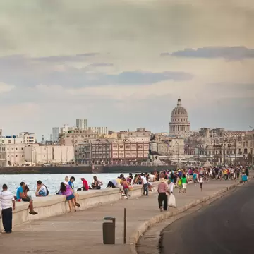 People sit along a sea wall in the evening as the sun starts to set. The city scape, dominated by a large domed building, stretches out behind them