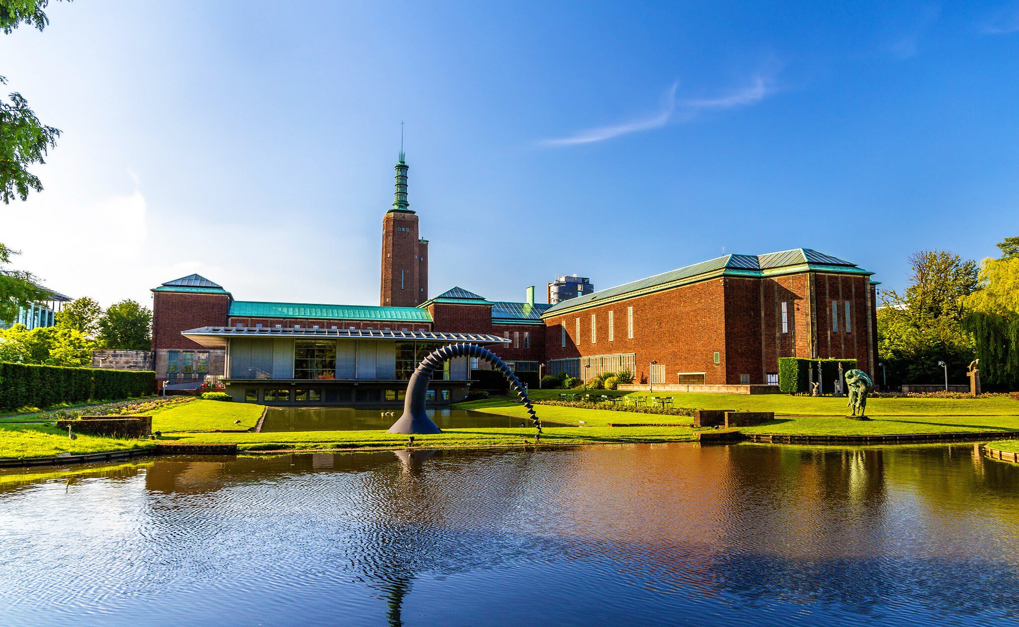 A brick museum with a spired tower and a sculpture beside a reflecting pond.