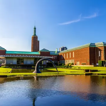 A brick museum with a spired tower and a sculpture beside a reflecting pond.