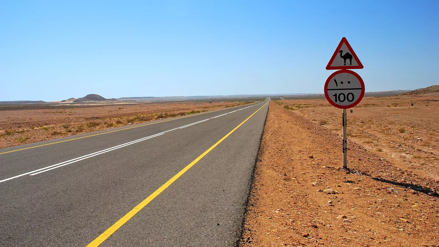 An empty paved road (with a camel crossing caution sign by the side) stretches out into the desert of Oman