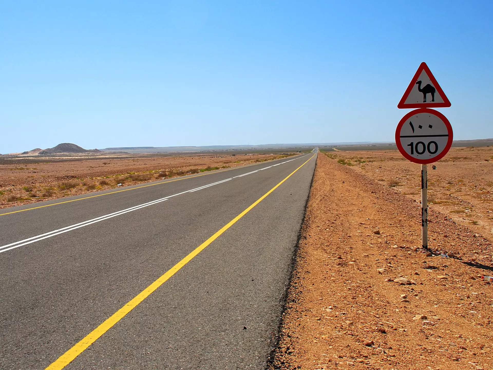 An empty paved road (with a camel crossing caution sign by the side) stretches out into the desert of Oman