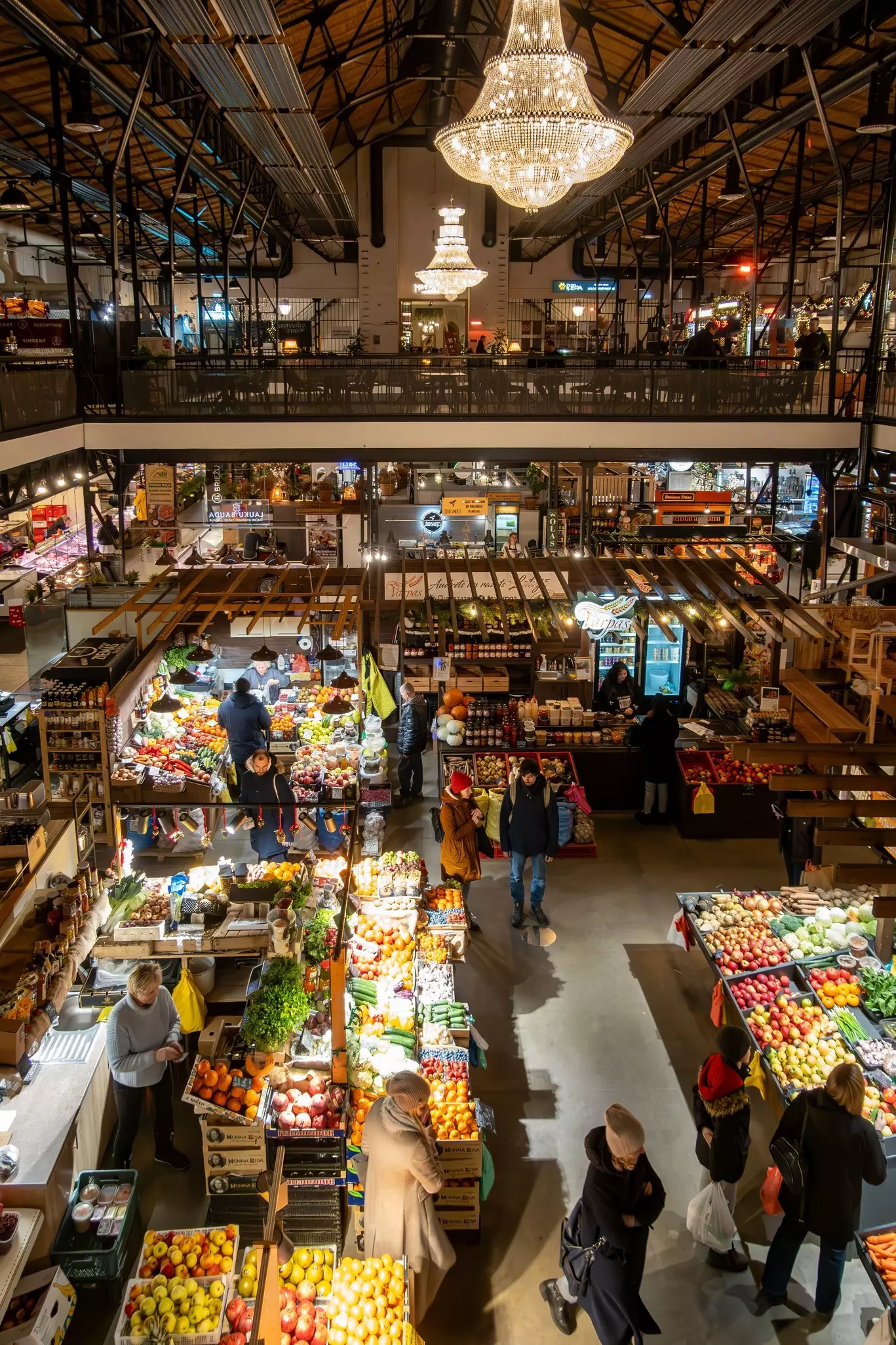 People shop for fresh fruit and vegetables in a covered food market with mezzanine seating for diners. Two large chandeliers hang from the market roof.