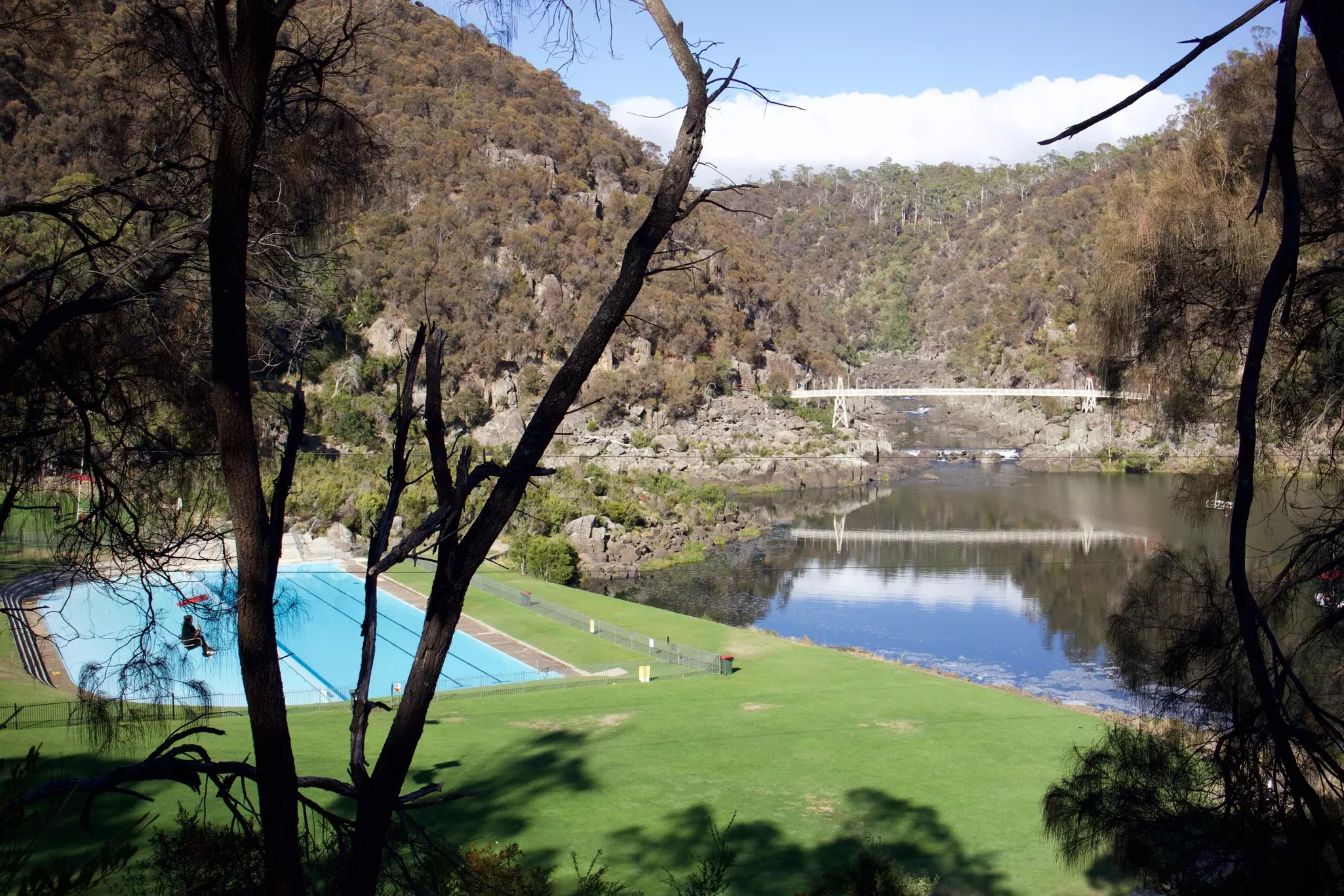 A scenic view of a grassy area featuring a swimming pool, surrounded by trees and a tranquil river. A bridge and hills are visible in the background, creating a serene landscape