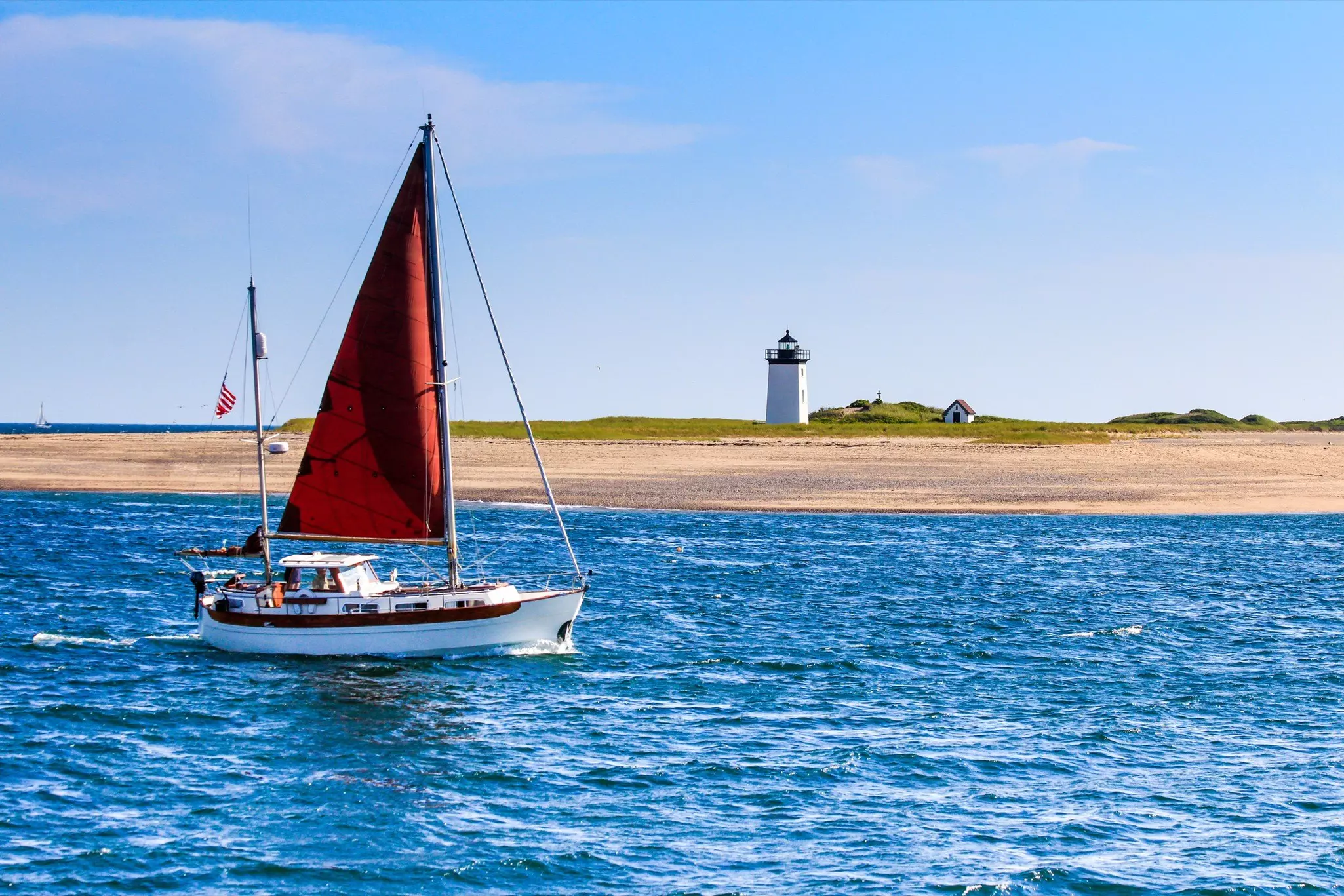 A sailboat with red sail hoisted travels on the sea past a white lighthouse and a little house with a red roof on the beach in the distance on a sunny day.