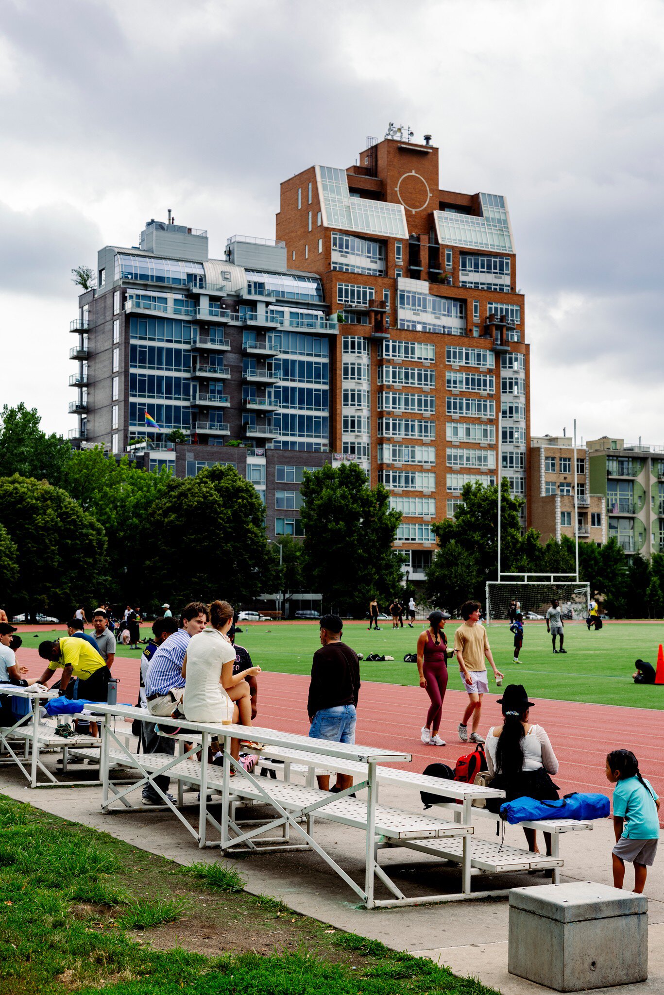 People on metal bleachers beside a red running track and field in McCarren Park, Brooklyn, below apartment towers.