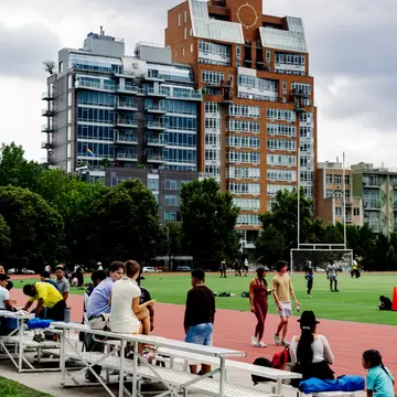 People on metal bleachers beside a red running track and field in McCarren Park, Brooklyn, below apartment towers.