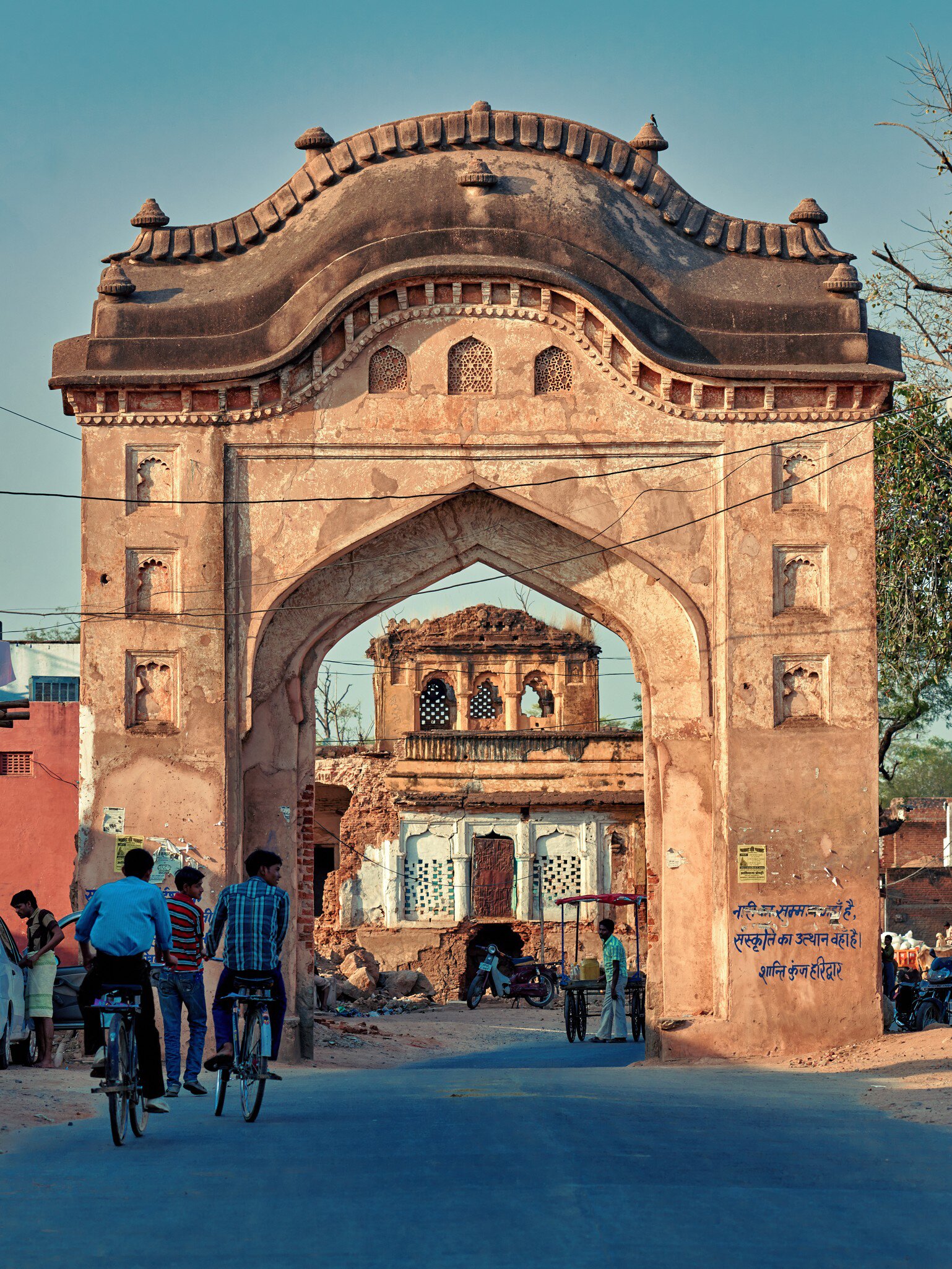 Weathered pink sandstone archway over a street, cyclists passing beneath and a ruined pavilion framed in the opening.