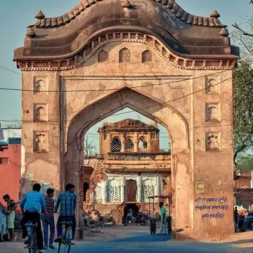 Weathered pink sandstone archway over a street, cyclists passing beneath and a ruined pavilion framed in the opening.