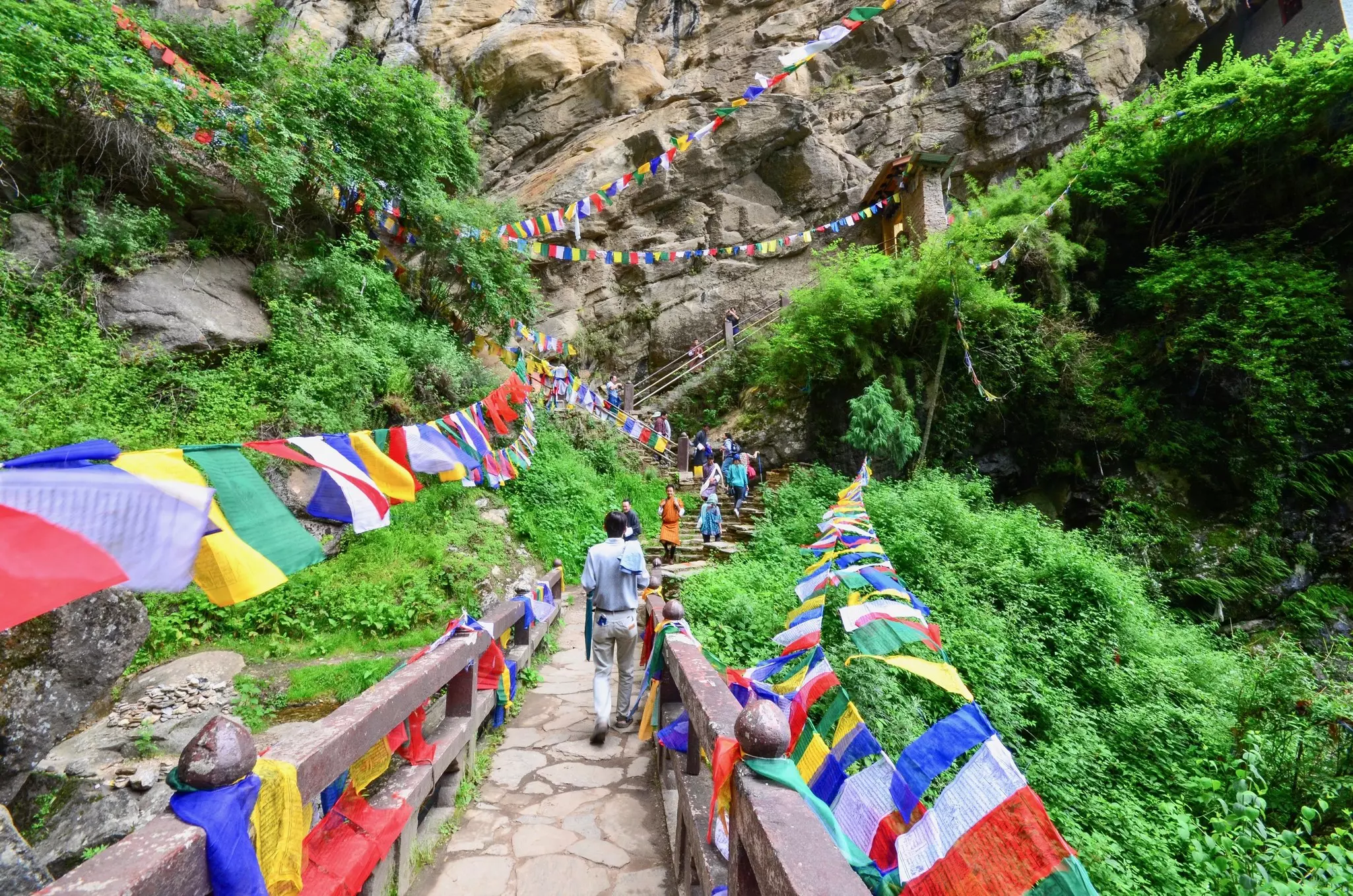 Pilgrims follow a path lined with prayer flags leading up to a series of stairs built into a rocky cliff face