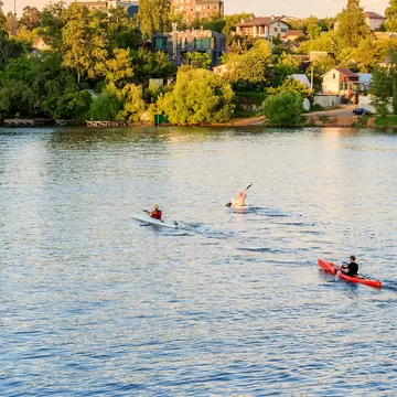 Three kayakers paddle across a sunlit river past trees and houses on the bank in Moscow