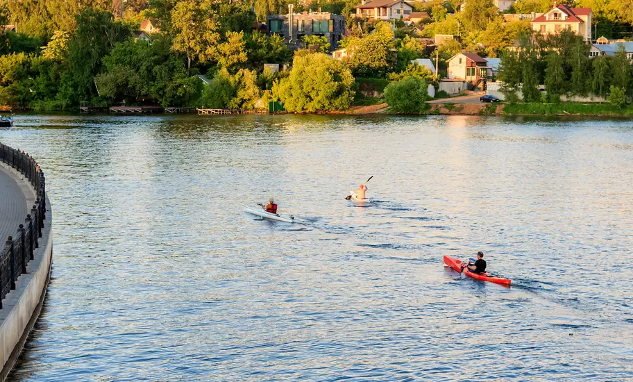 Three kayakers paddle across a sunlit river past trees and houses on the bank in Moscow