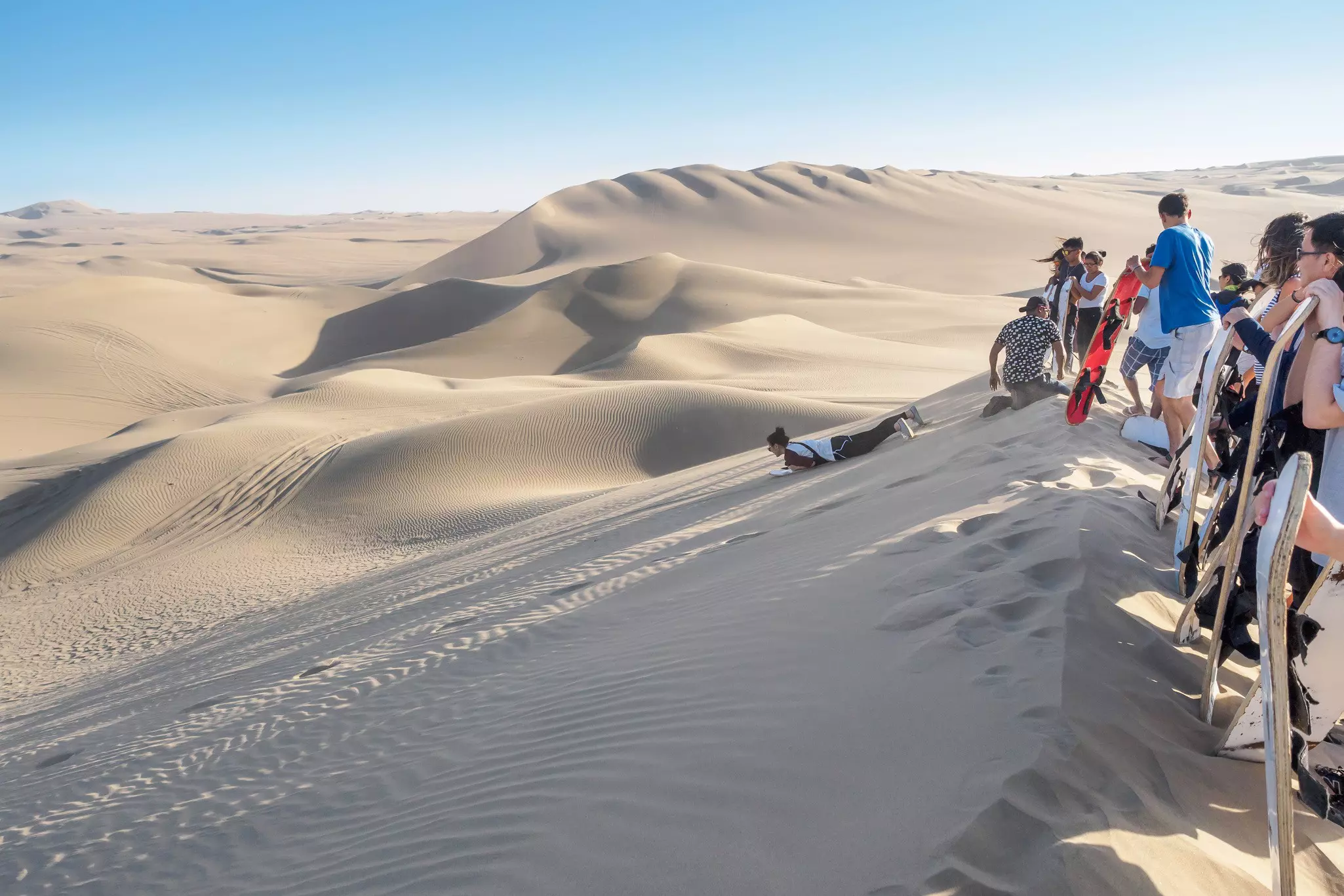 Tourists stand at the ridge of a massive sand dune as they wait to go sandboarding in the desert