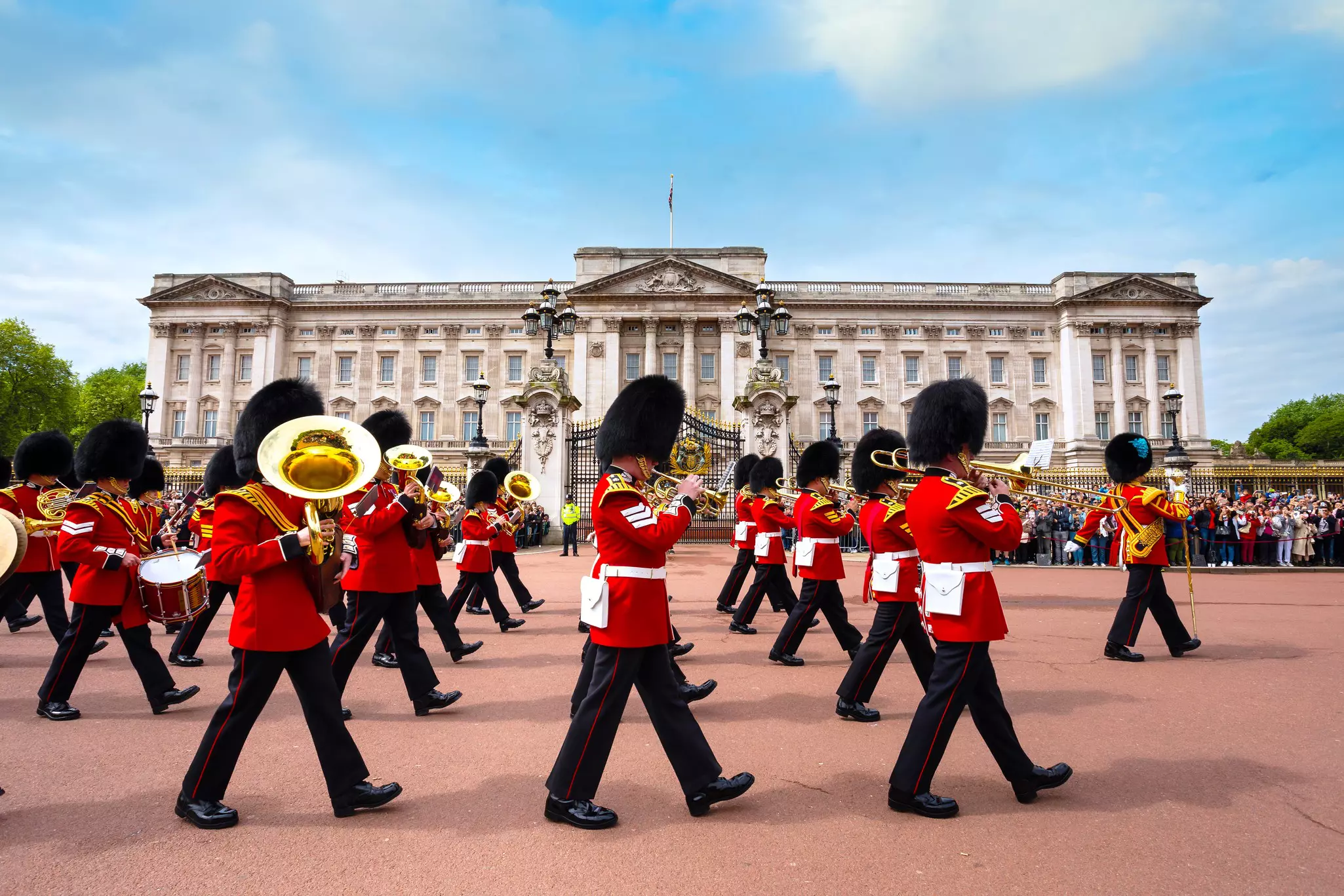 A miltary band playing brass wind instruments parades past a vast palace building with crowds of onlookers.