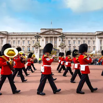 A miltary band playing brass wind instruments parades past a vast palace building with crowds of onlookers.