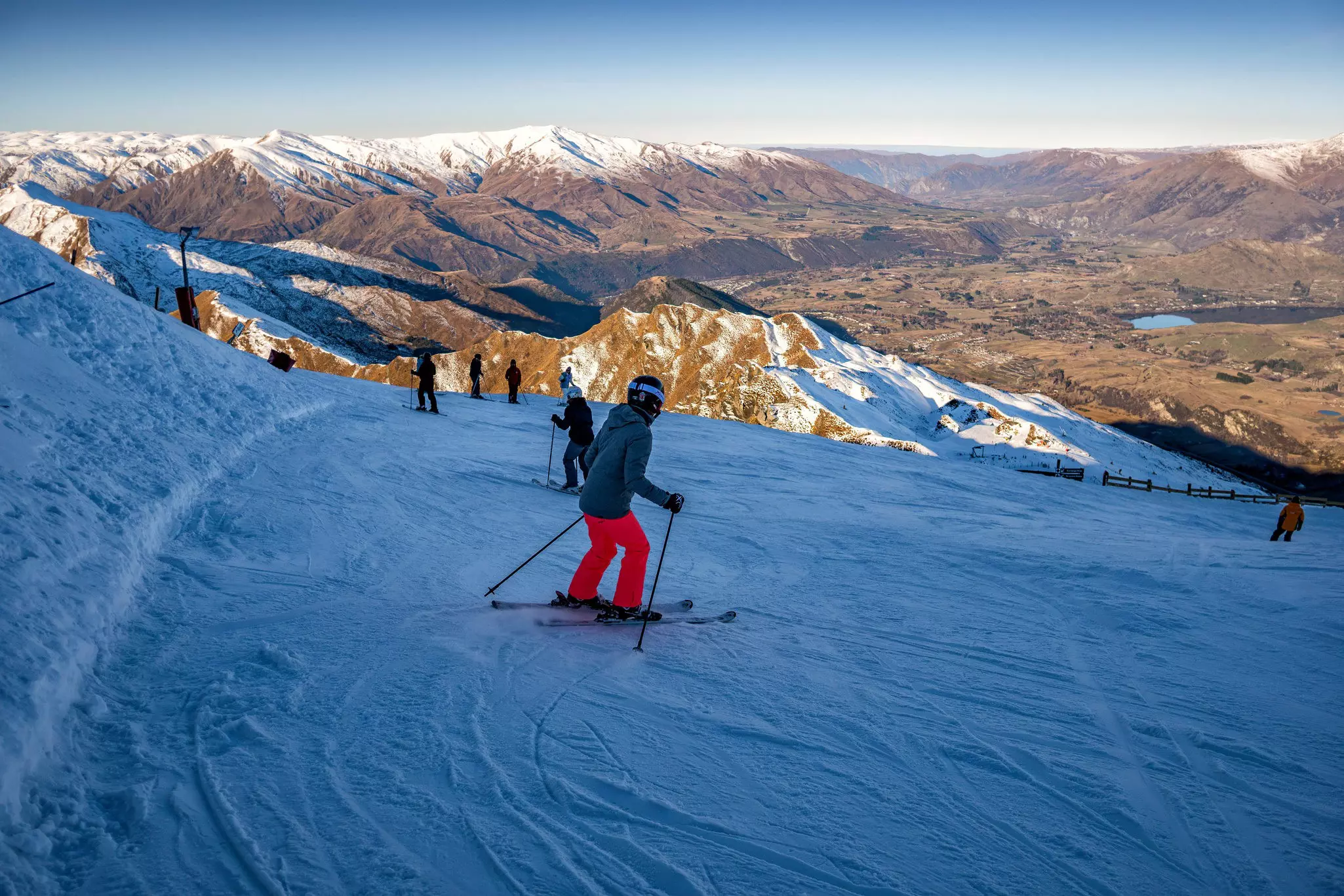 A skier in winter gear and a helment heads down a snowy slope as the setting sun casts long shadows over the valley.