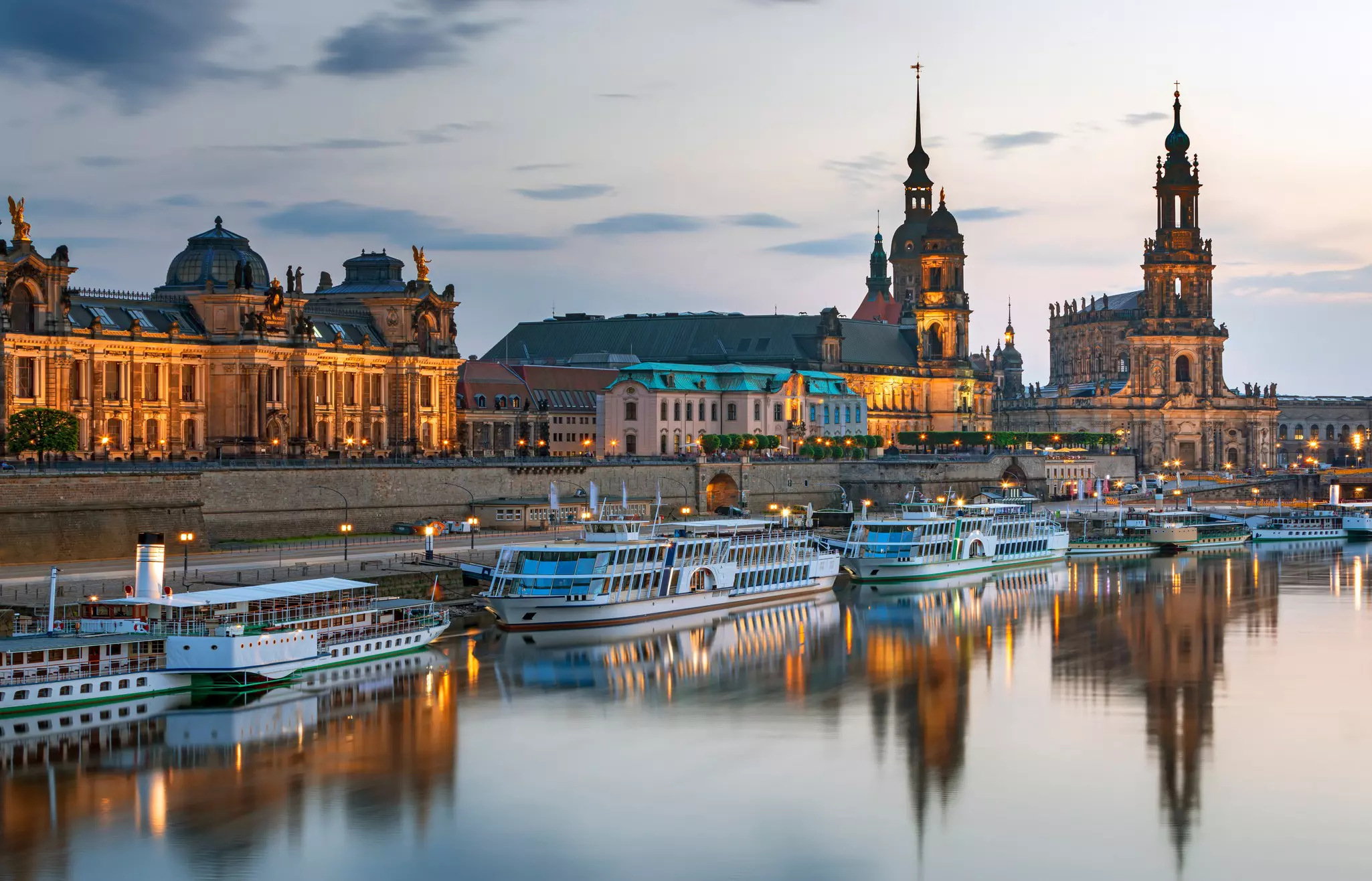 A series of riverboats are moored along a river in a city at dusk. Historic buildings lining the river are illuminated.